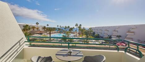 A balcony with two chairs and a table, overlooking a pool area with palm trees and a clear sky.