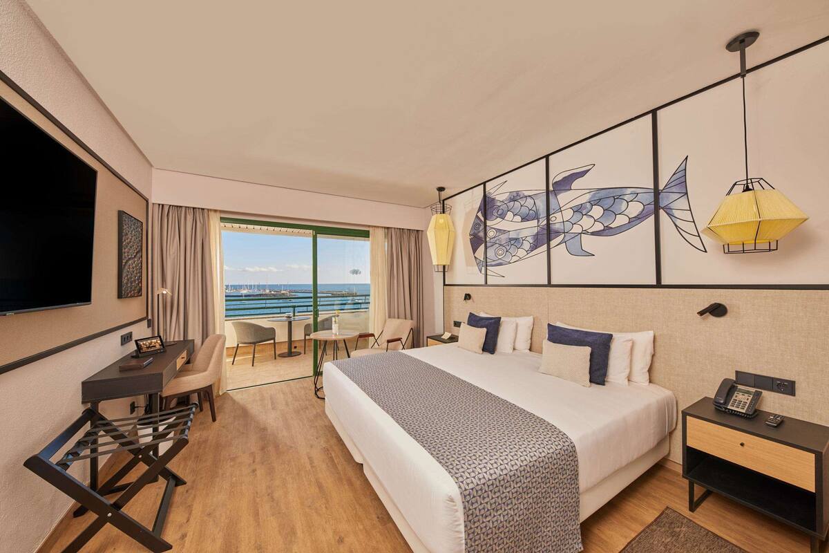 A hotel room with a large bed, a desk, a chair, and a view of the sea.