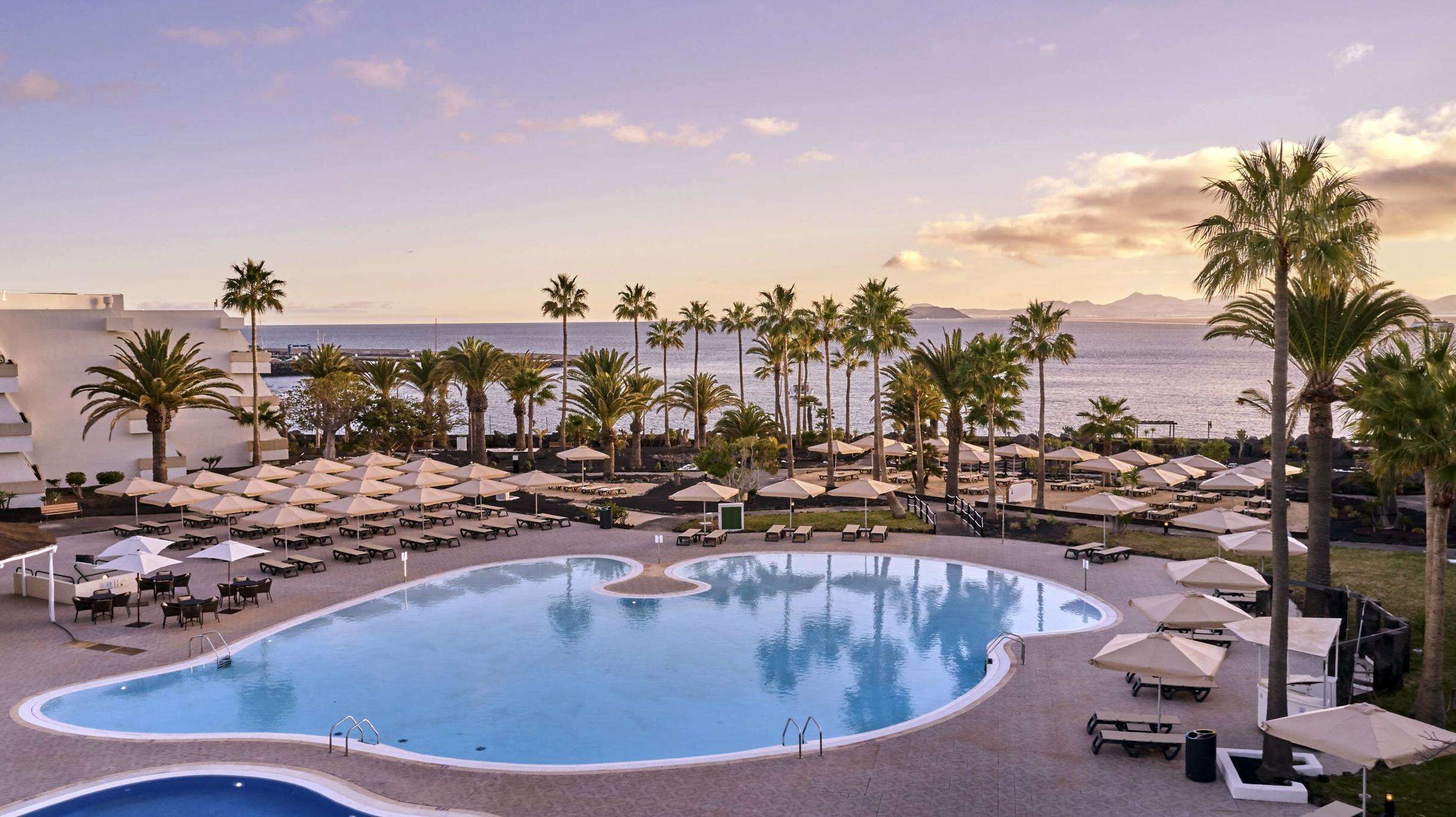 A resort pool area with sun loungers and umbrellas, surrounded by palm trees and a view of the sea.