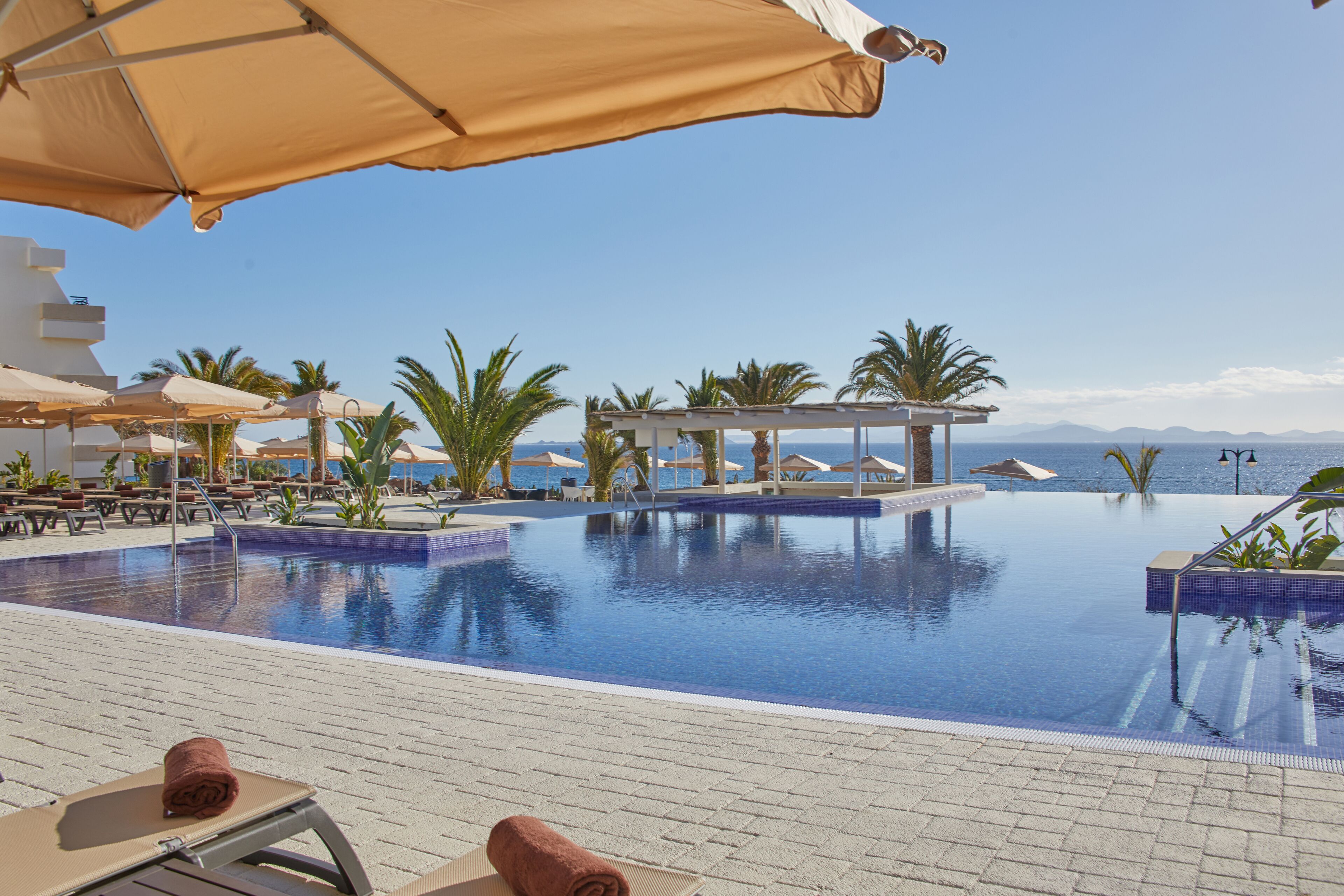 Zona de piscina con sillones de descanso y sombrillas, rodeada de palmeras y vista despejada al mar.
