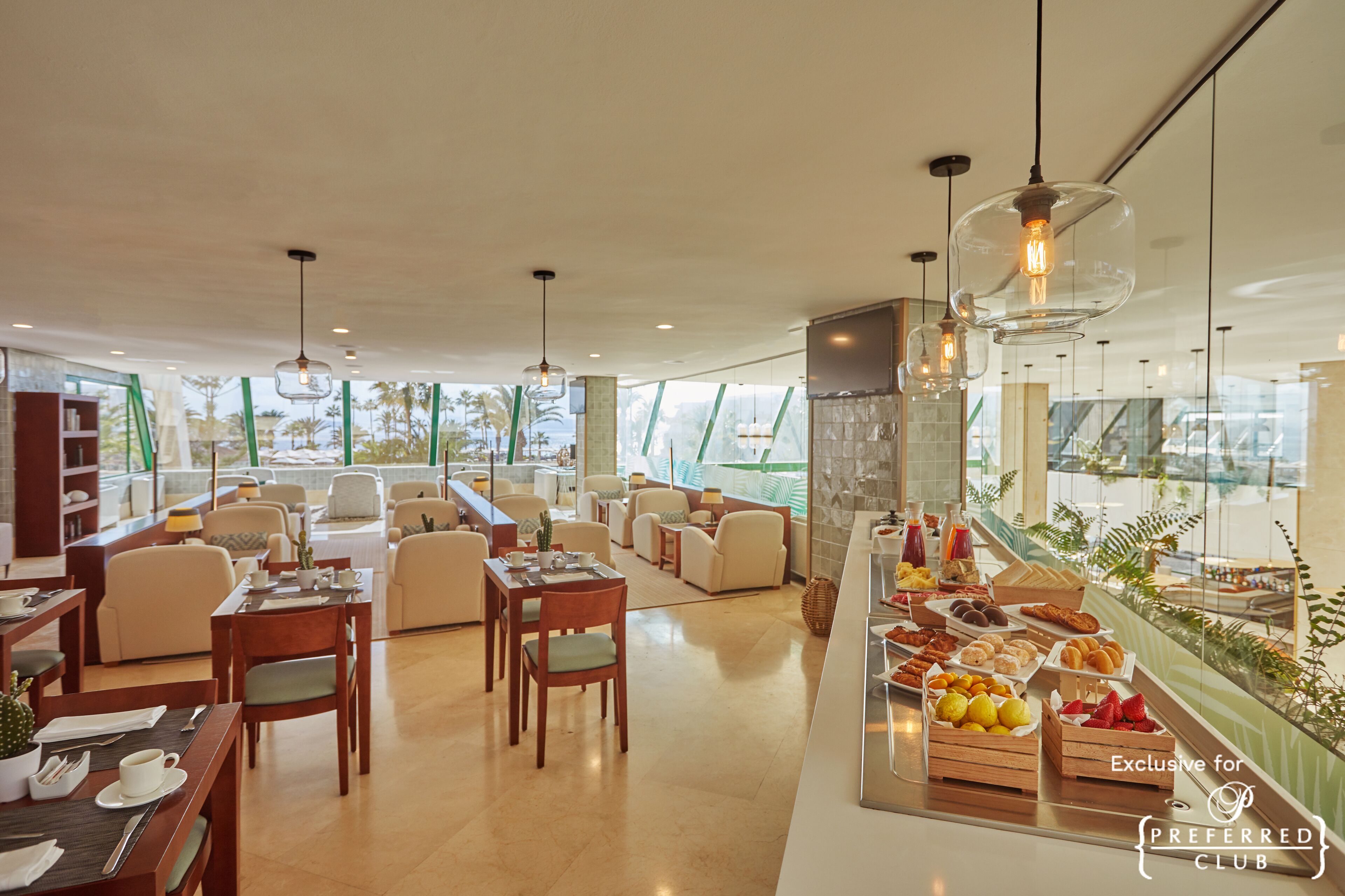 A spacious hotel lobby with a dining area, comfortable seating, and a buffet with fresh fruits and pastries.