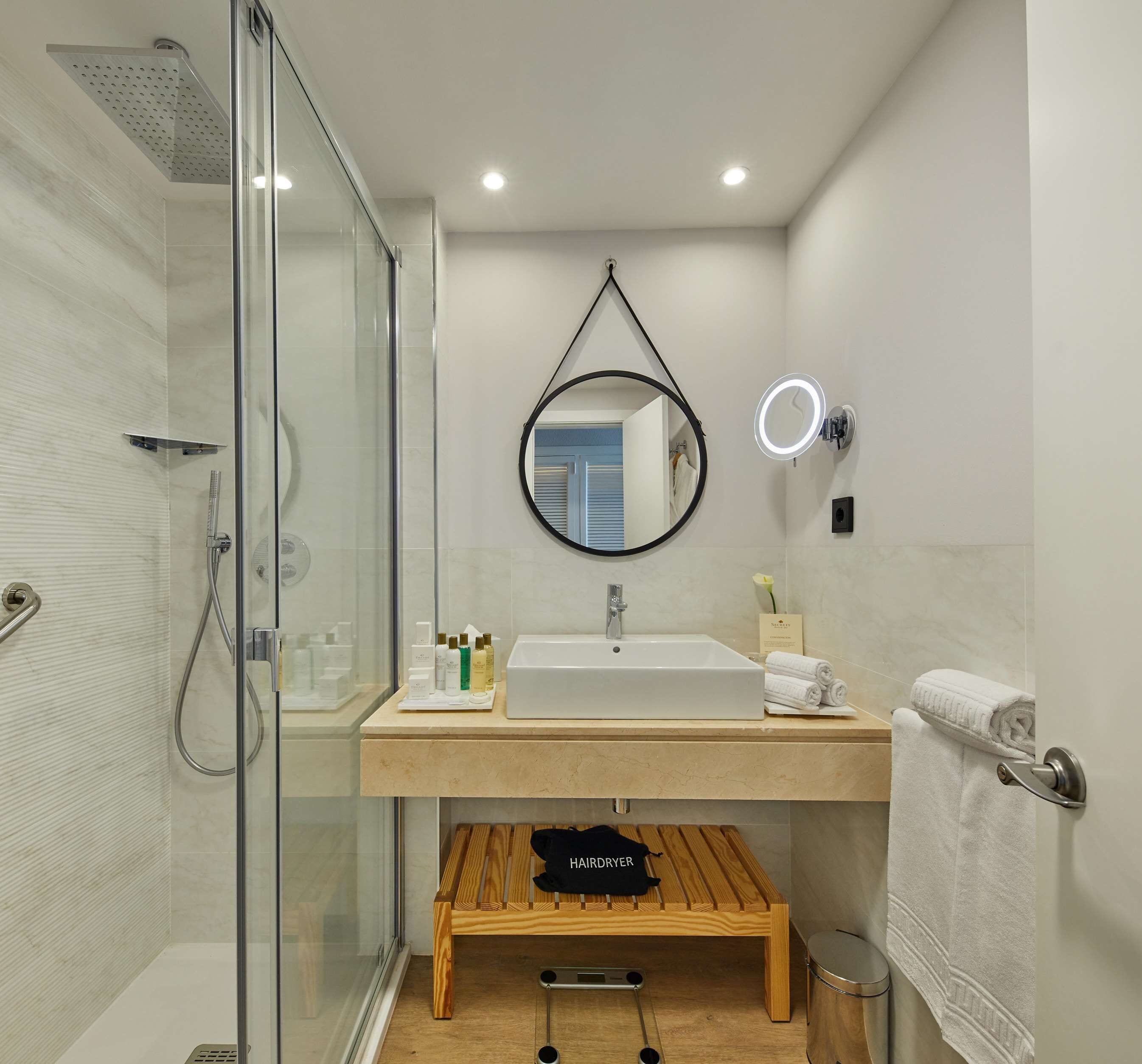 Een moderne badkamer met een glazen douchecabine, een wastafel met spiegel erboven en een houten bankje.
