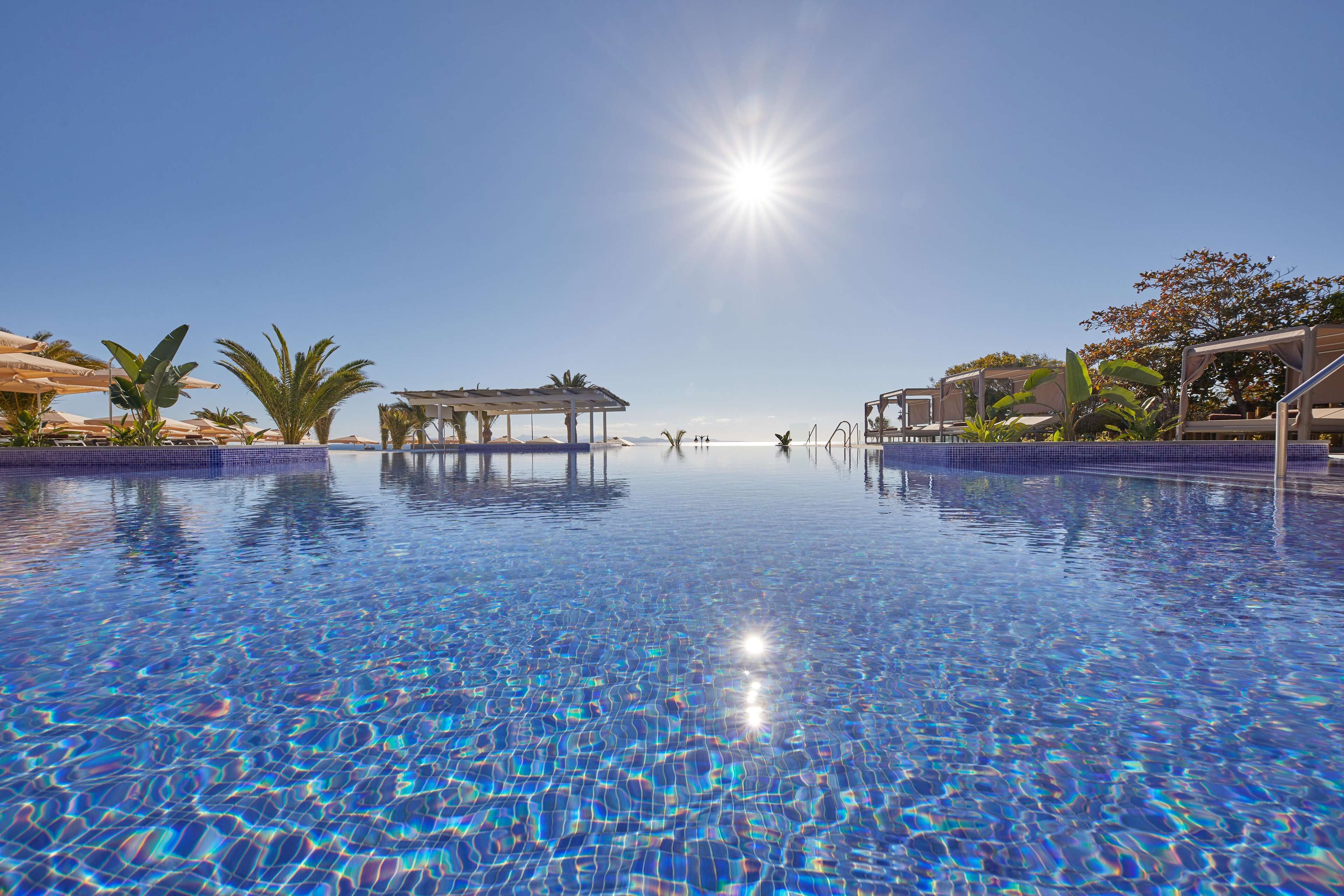 Piscina azul cristalina con reflejos de sol, rodeada de palmeras y una zona cubierta.