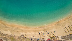 Vista aérea de una playa con agua turquesa cristalina, orilla de arena y filas de tumbonas.