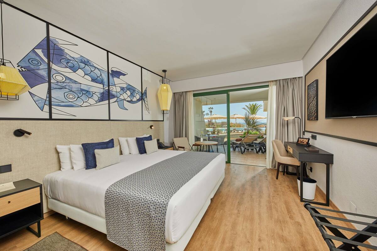 A hotel room with a large bed, a desk, a chair, a TV, and a balcony with outdoor seating.