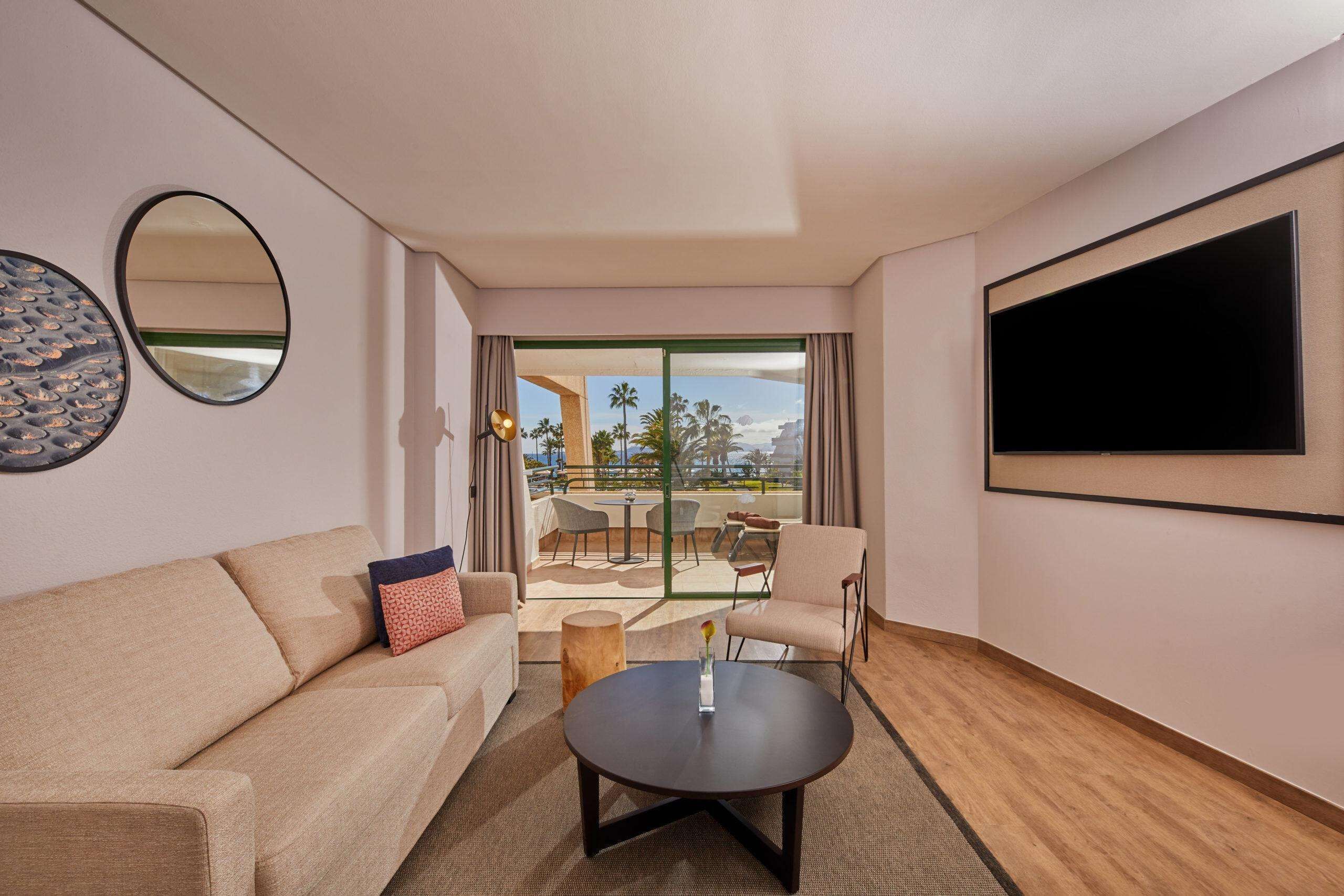 Una habitación de hotel moderna con sofá, mesa de centro, televisor y balcón con mobiliario exterior.