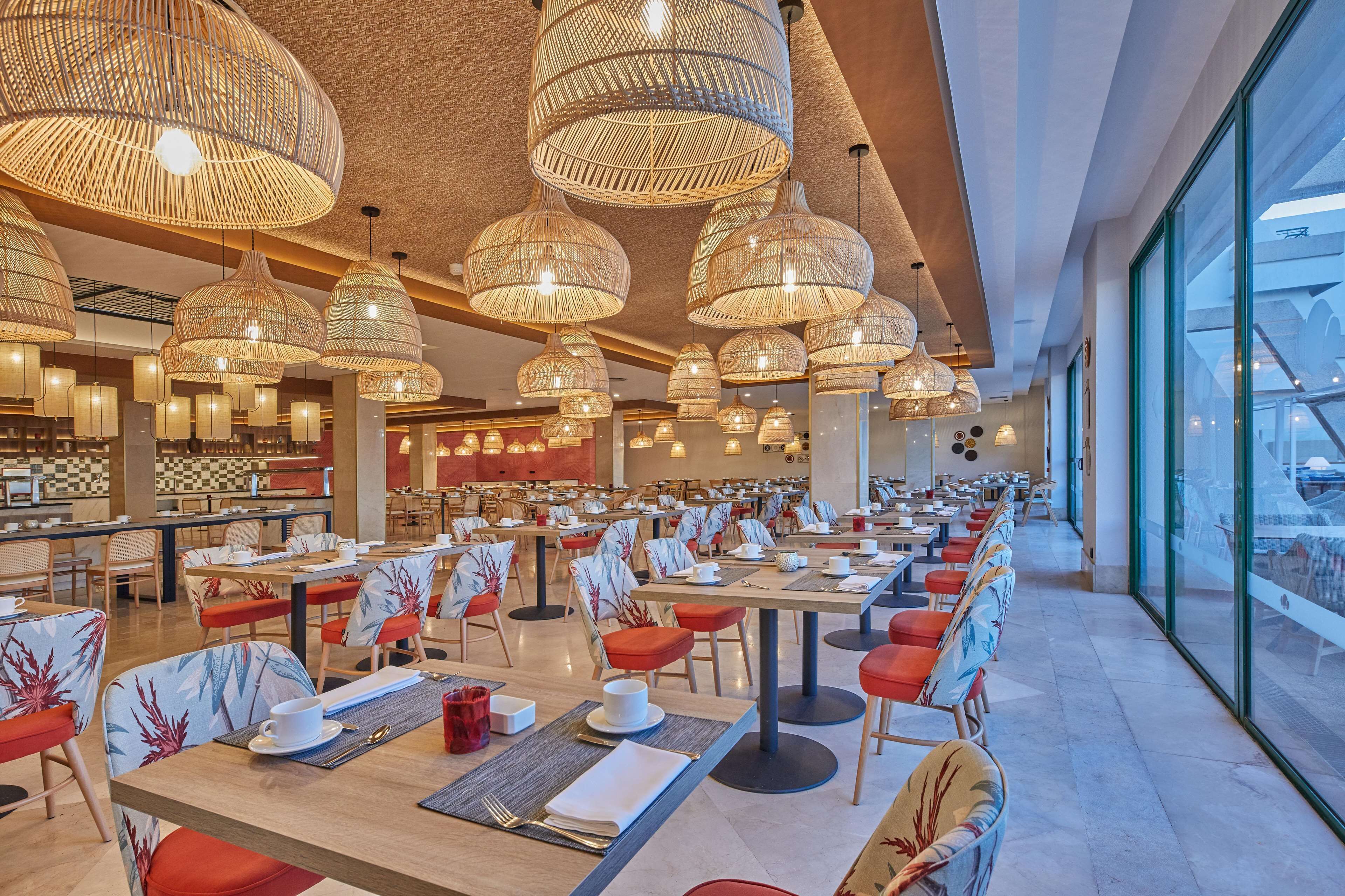 A spacious dining area with multiple tables and chairs, large hanging wicker chandeliers, and a view of the outside through large windows.