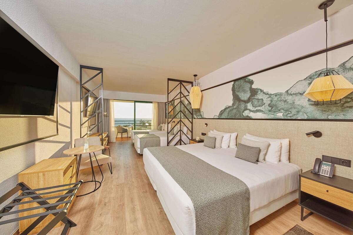 A modern hotel room with a large bed, a desk, and a chair. There is a painting on the wall and a window with a view.