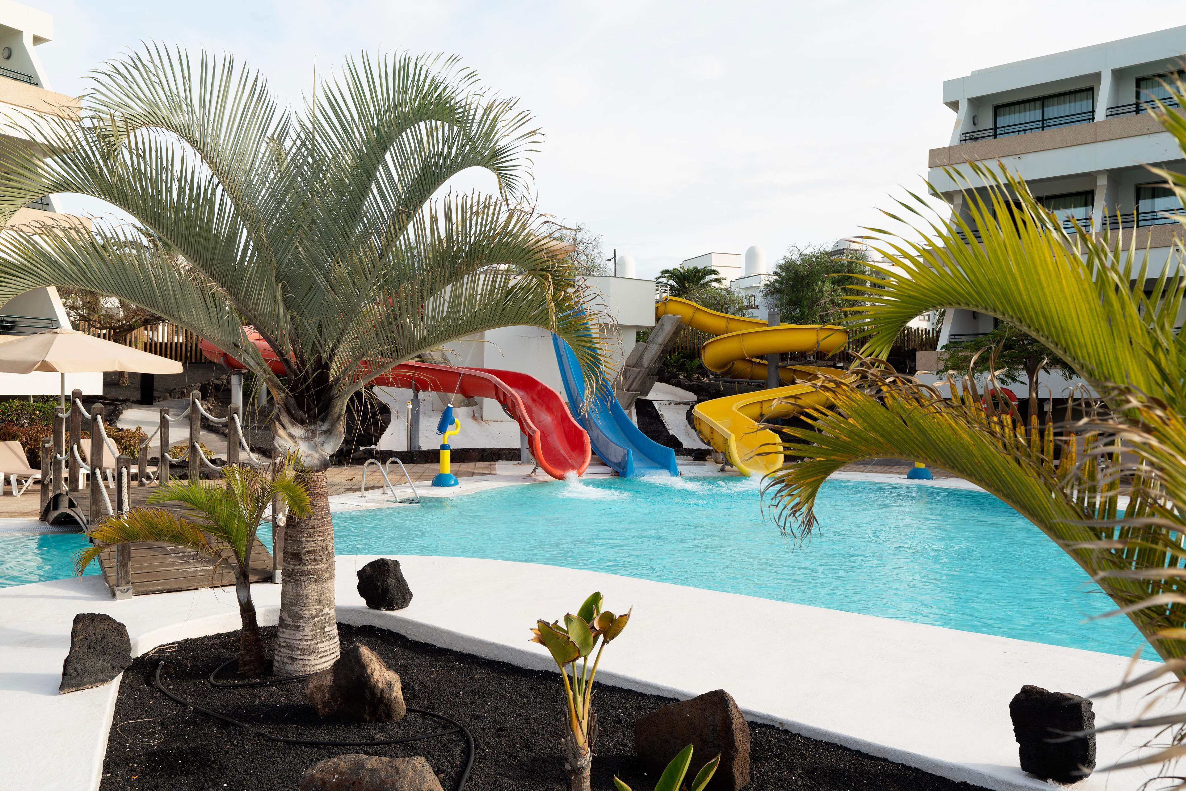 Zona de piscina con varias toboganes, palmeras y un edificio al fondo.