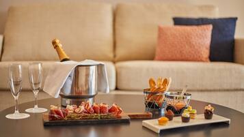 A dining table with a bottle of champagne, two glasses, a basket of bread, and a variety of pastries.