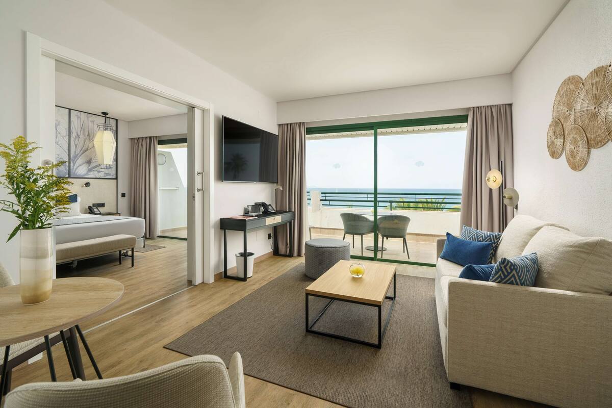 A modern hotel room with a sofa, a dining table, a TV, and a balcony with a view of the sea.