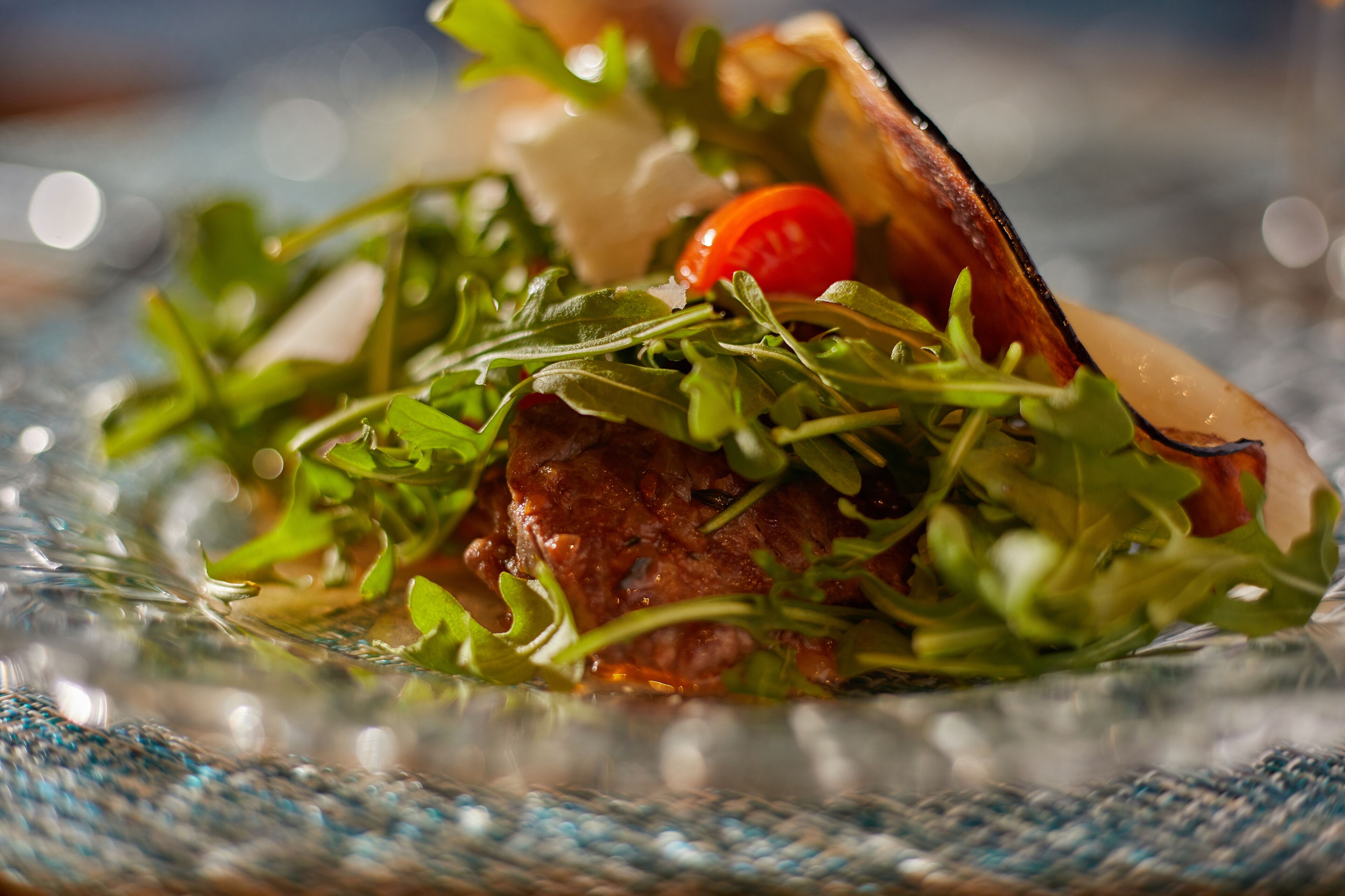 A close-up of a dish with greens, a tomato, and a piece of meat, served on a glass plate.