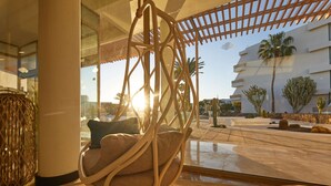 A hanging chair with cushions in a modern interior with large windows, a view of palm trees, and a pool area.