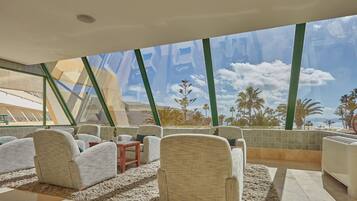 Un salón moderno con asientos cómodos y un ventanal que ofrece vistas a palmeras y una playa.