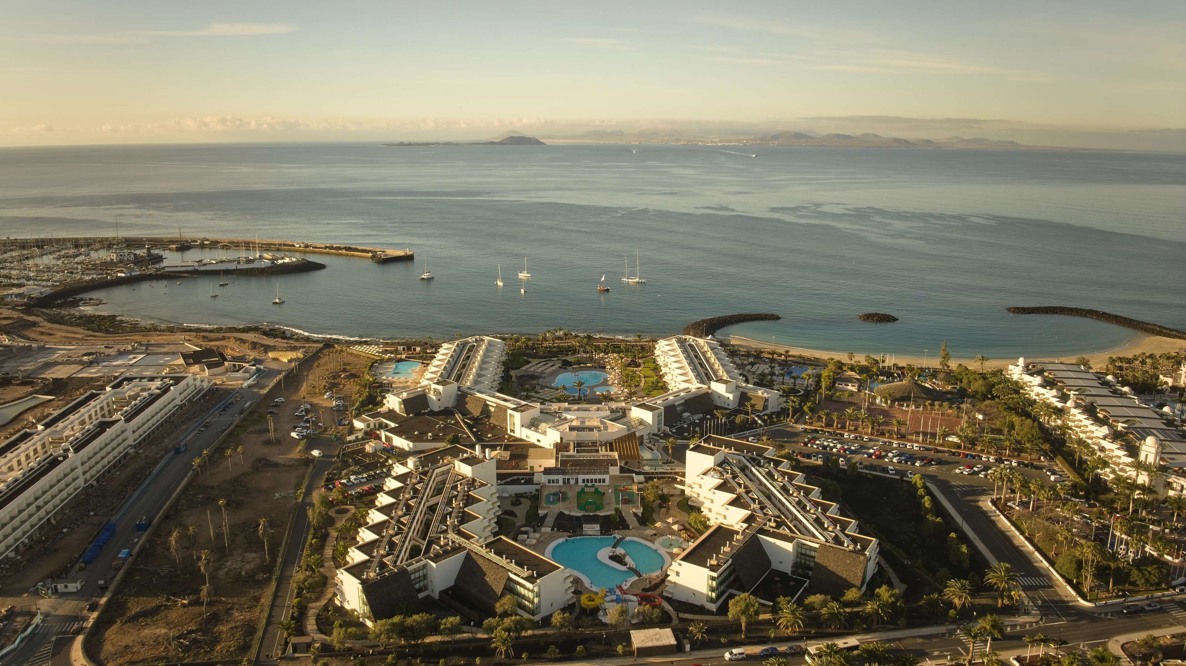 An aerial view of a coastal resort with multiple swimming pools, a marina with boats, and a beach area.