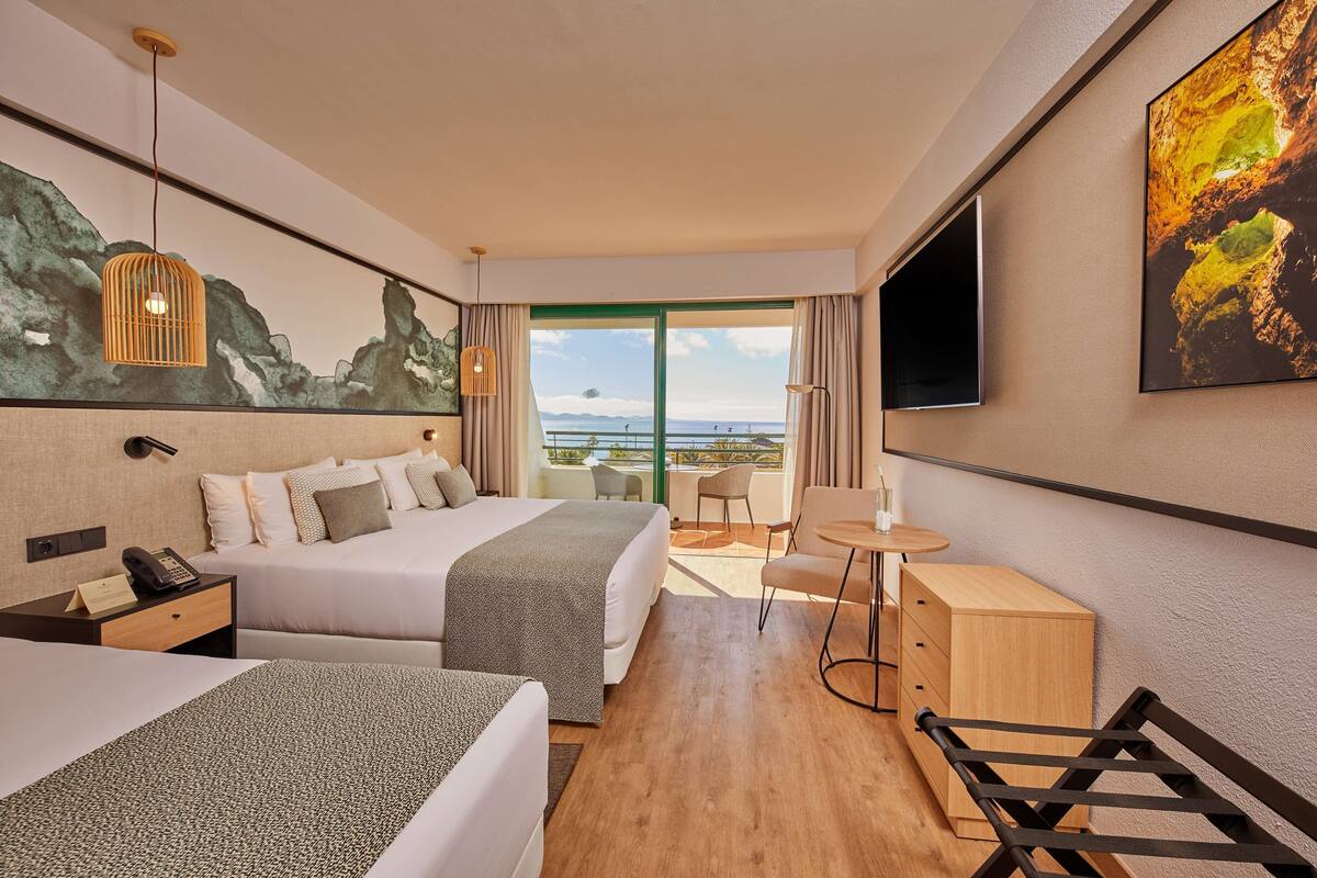 A hotel room with two beds, a desk, a chair, a small table, a TV, and a large window with a view of the sea.