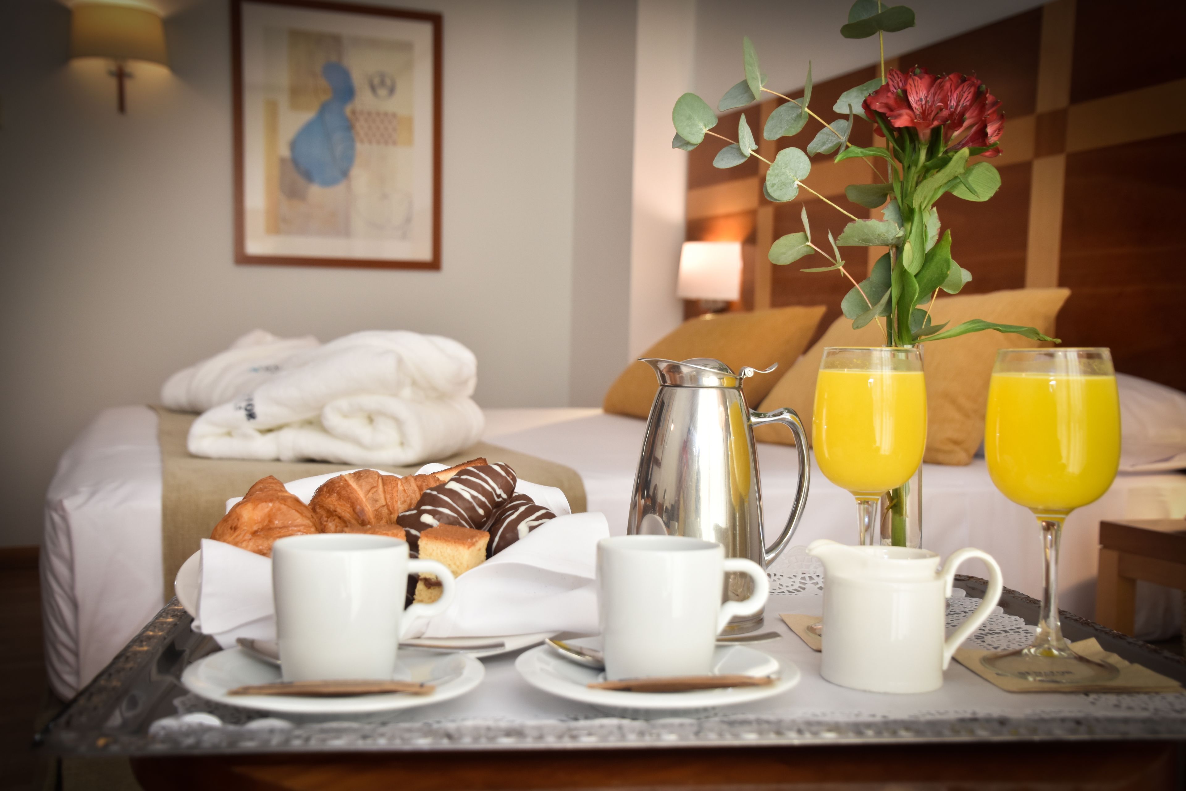 Un vassoio per la colazione con croissant, una caffettiera e due bicchieri di succo d'arancia su un letto con un cuscino e una coperta.