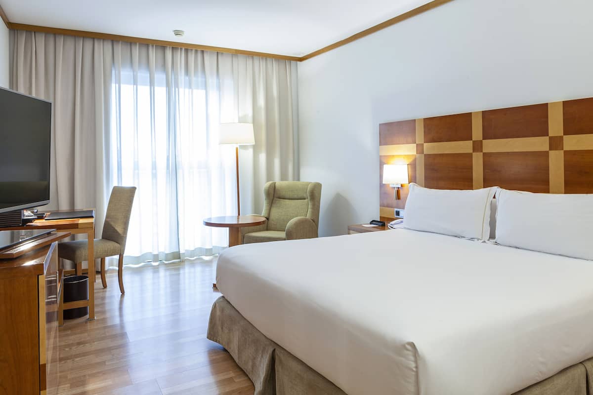 Une chambre d’hôtel avec un grand lit, un bureau avec une télévision, une chaise, une lampe et une vue sur l’extérieur à travers des rideaux transparents.