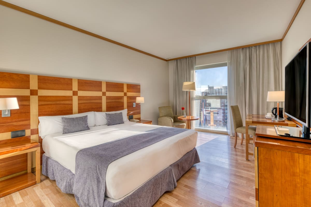 Une chambre d’hôtel dotée d’un grand lit, d’une tête de lit en bois, d’un bureau avec une télévision et offrant une vue sur la ville grâce à de grandes fenêtres.