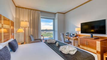 A hotel room with a large bed, a flat-screen TV, a desk with a chair, a lamp, and a view of the city.