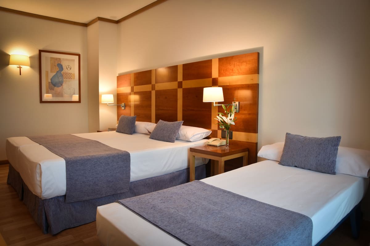 Une chambre d’hôtel avec deux lits, chacun recouvert d’une couverture grise et agrémenté d’oreillers, dotée d’une tête de lit en bois, de lampes de chevet et d’une œuvre d’art encadrée au mur.