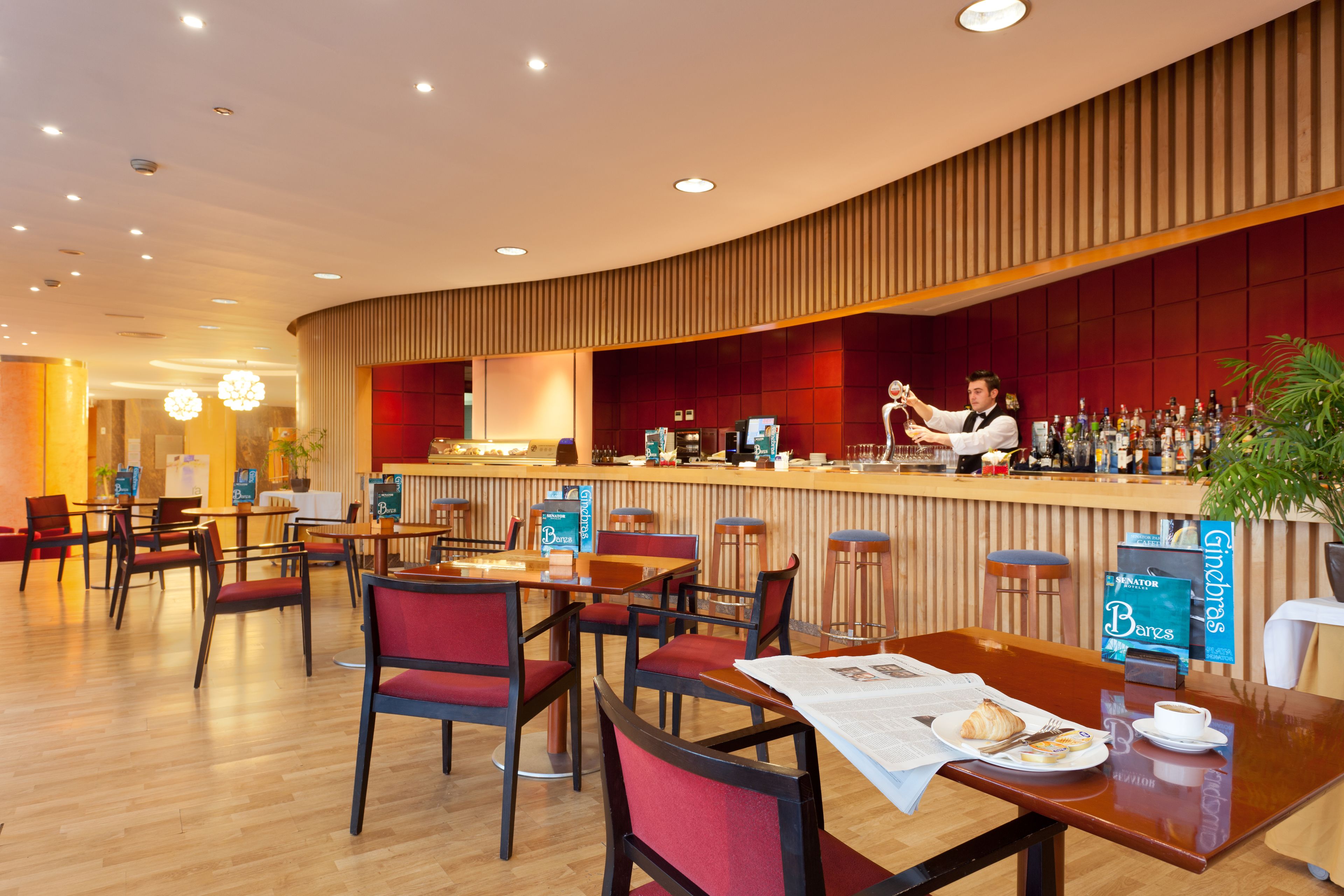 A modern restaurant with a curved wooden bar, red upholstered chairs, and a breakfast setting with croissants and coffee.