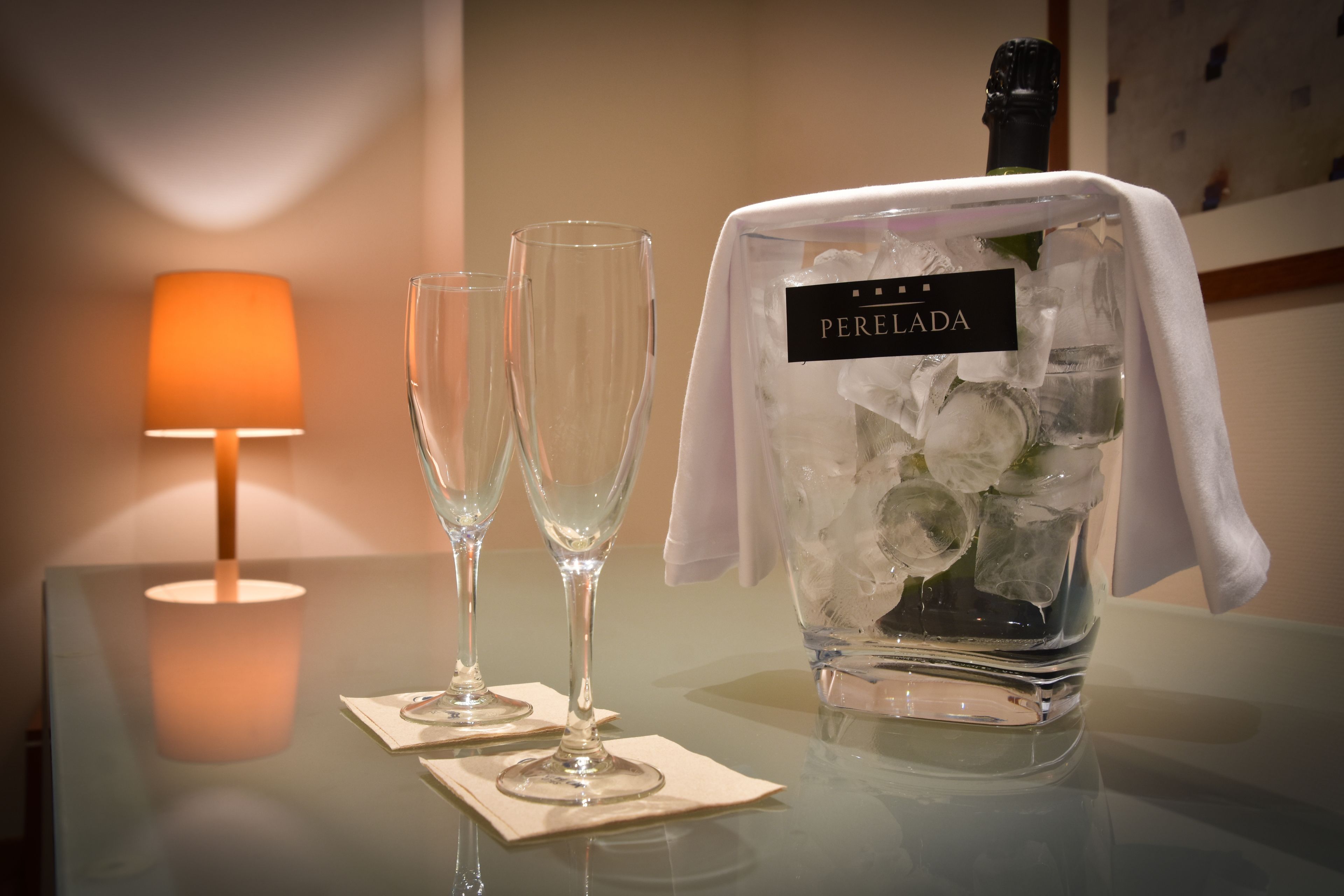 Une bouteille de champagne PereLada avec des glaçons, deux coupes à champagne vides et une lampe de table allumée, le tout posé sur une surface réfléchissante.