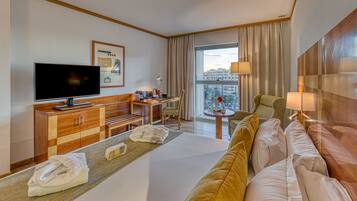 Habitación de hotel con una cama grande, un televisor, un escritorio con una silla, una mesita con una lámpara y vista a la ciudad desde la ventana.