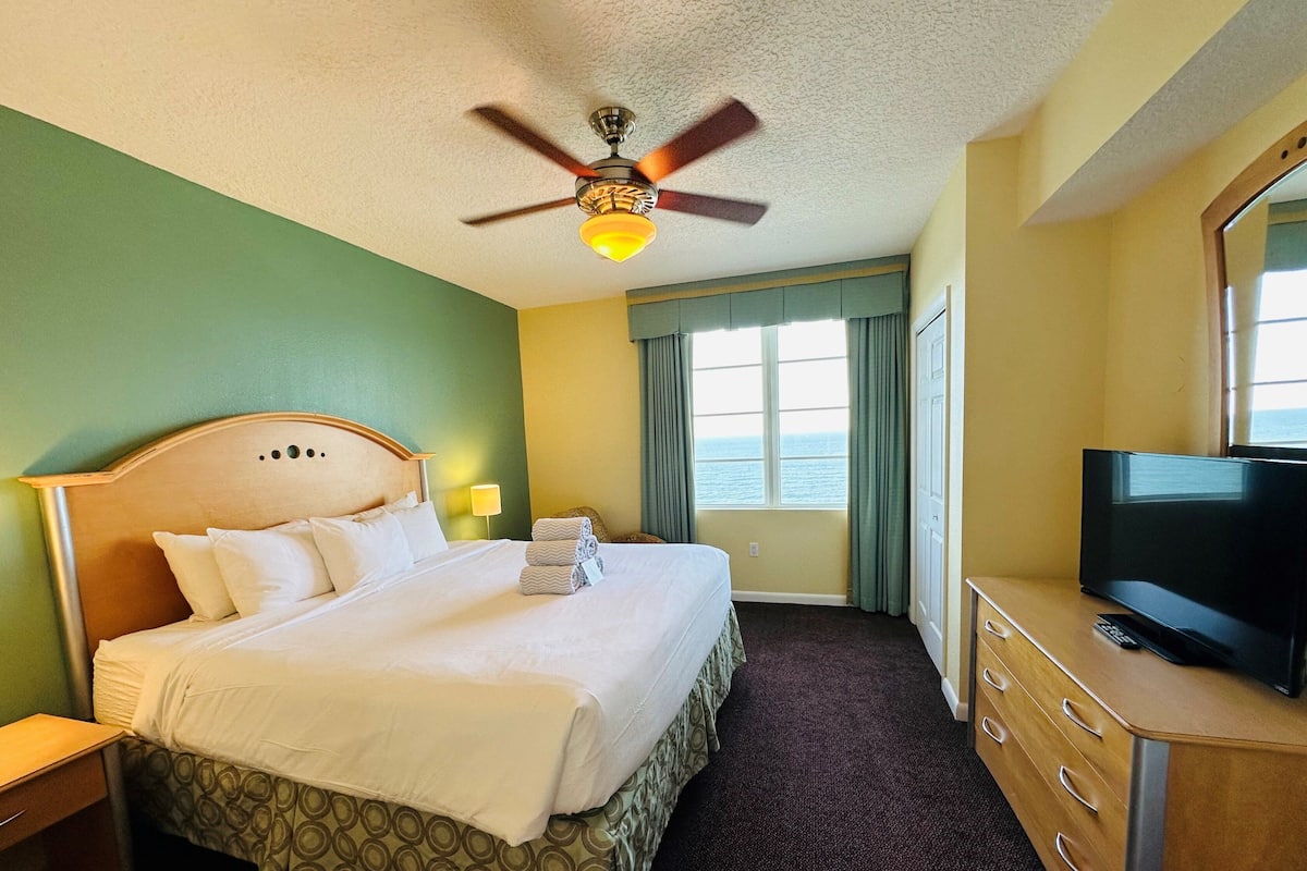 A hotel room with a bed, a television, a ceiling fan, a window with a view of the sea, and a dresser.