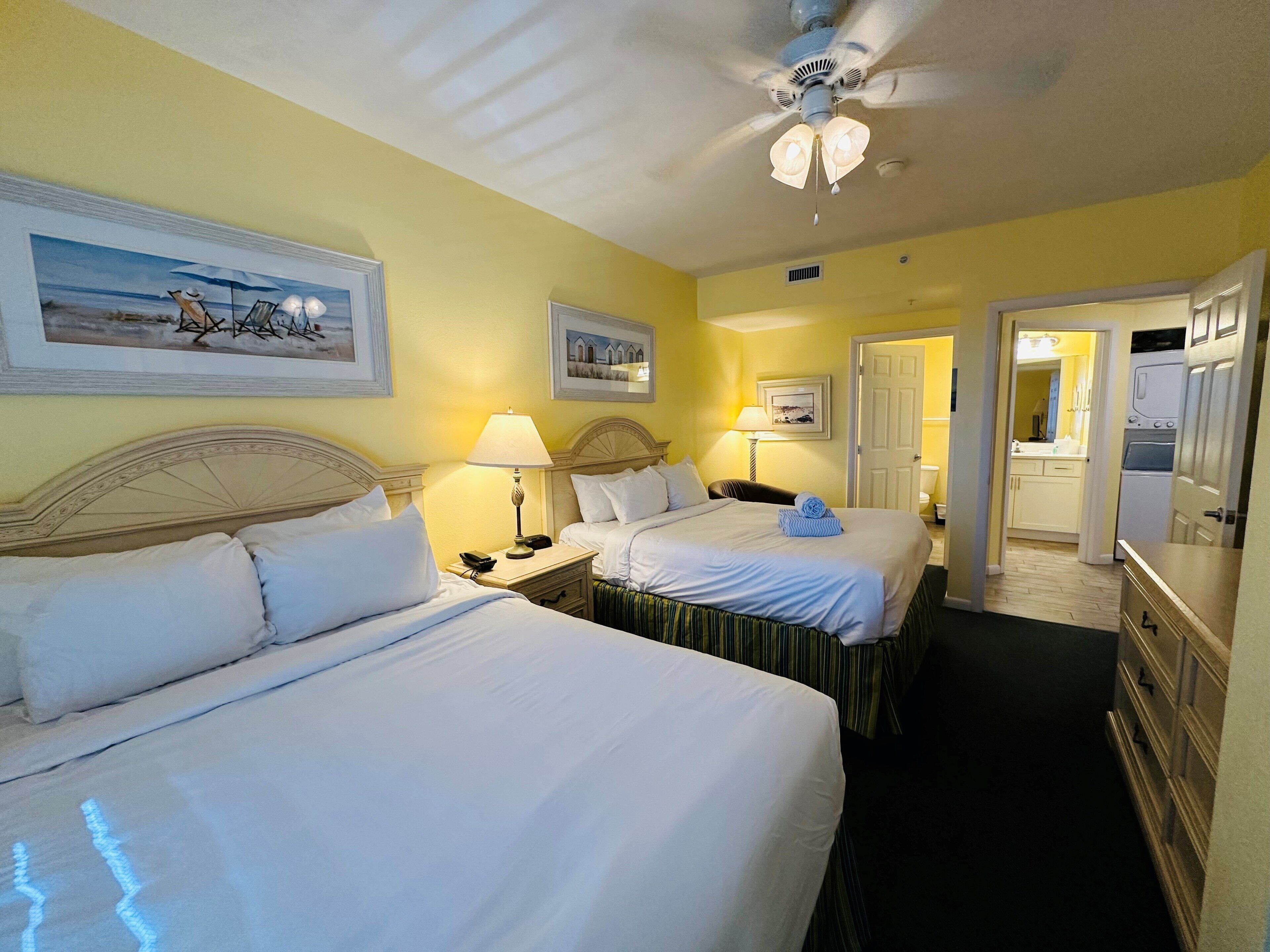 A hotel room with two beds, a ceiling fan, and artwork on the walls.