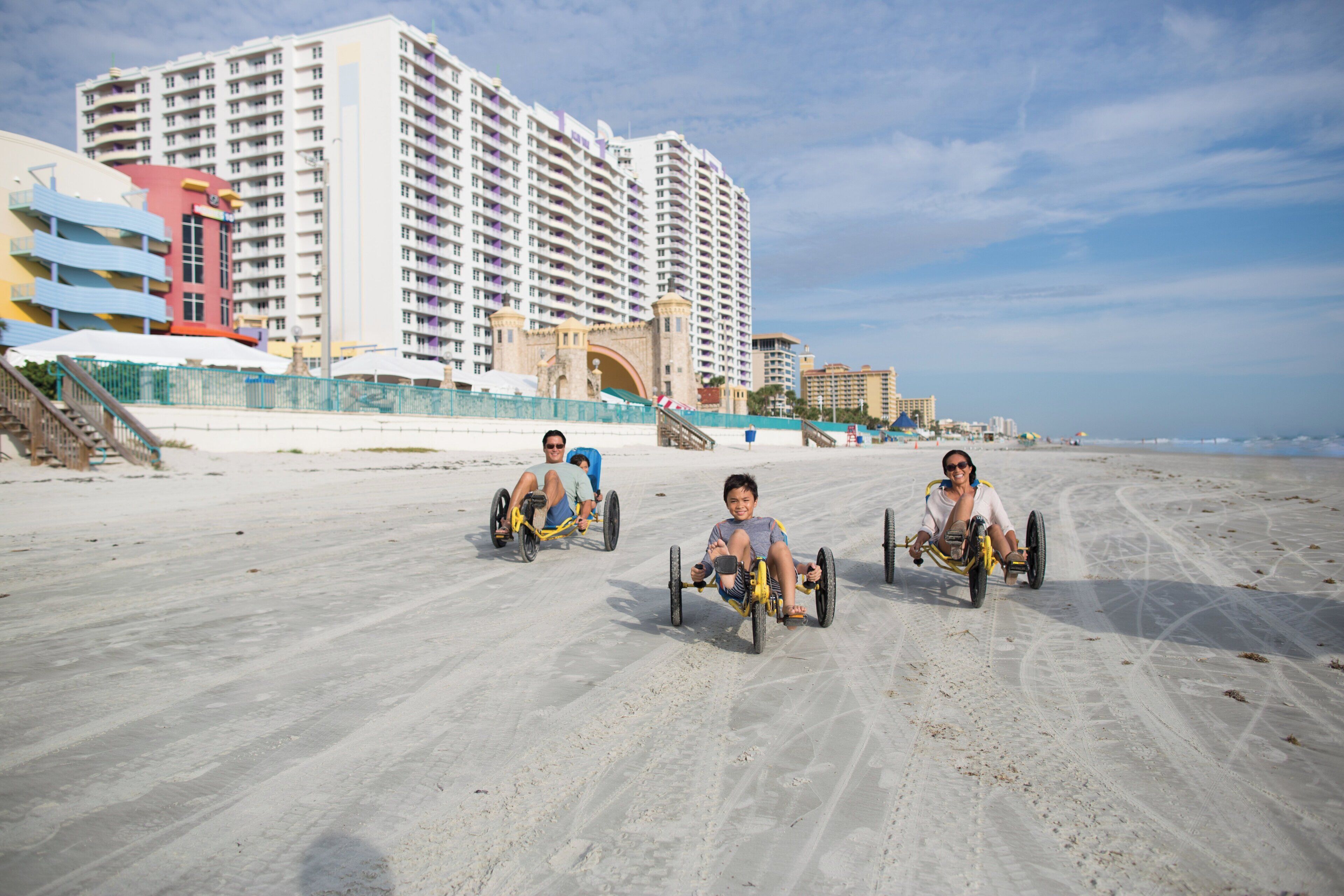 Une plage de sable où l’on voit trois personnes sur des tricycles. Au fond, on aperçoit des immeubles à plusieurs étages dotés de balcons.