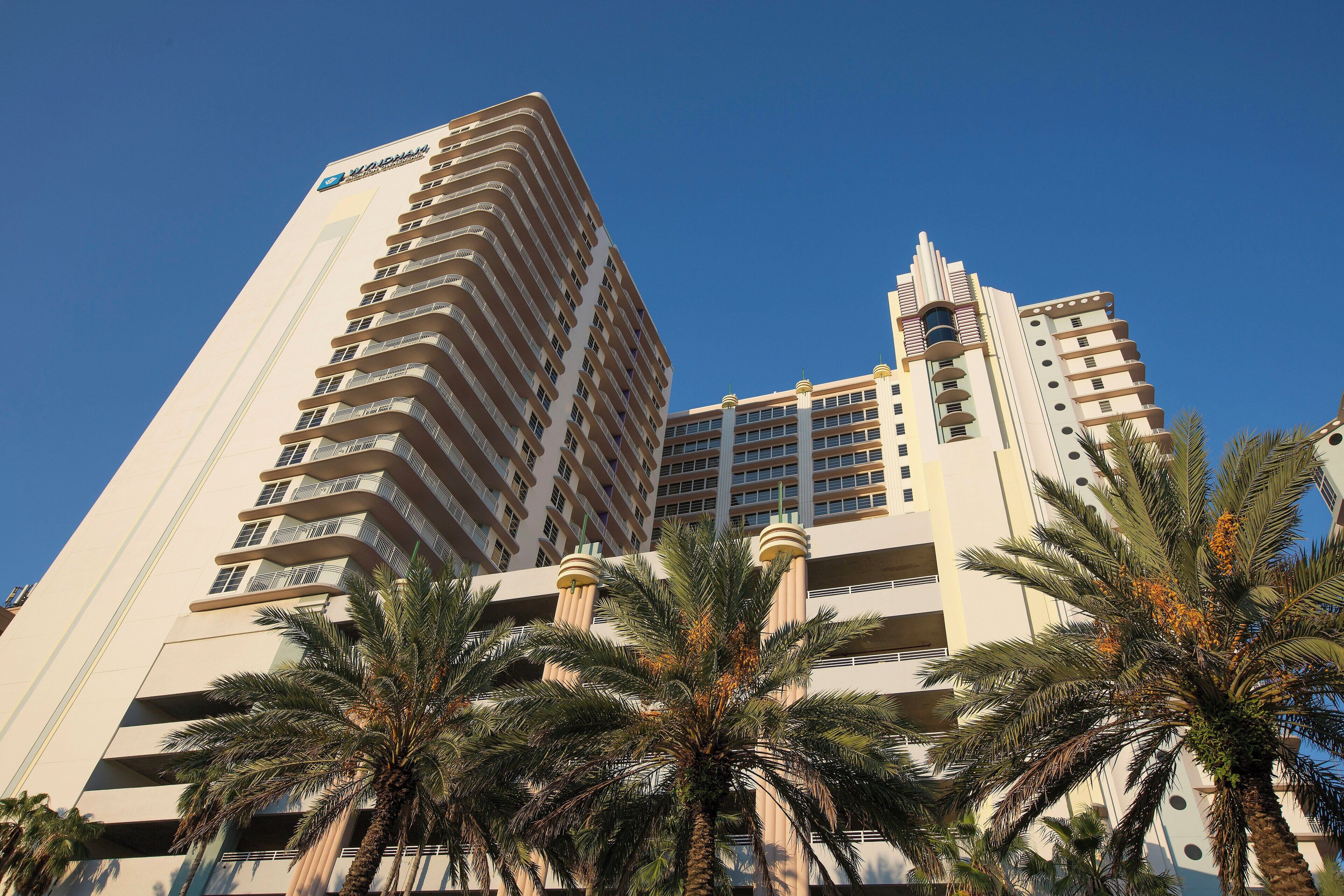 A high-rise hotel with multiple balconies, palm trees in the foreground, and a clear blue sky.