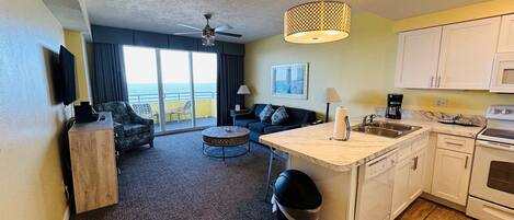 A hotel room with a balcony, a kitchenette, a living area with a sofa and armchairs, and a ceiling fan.