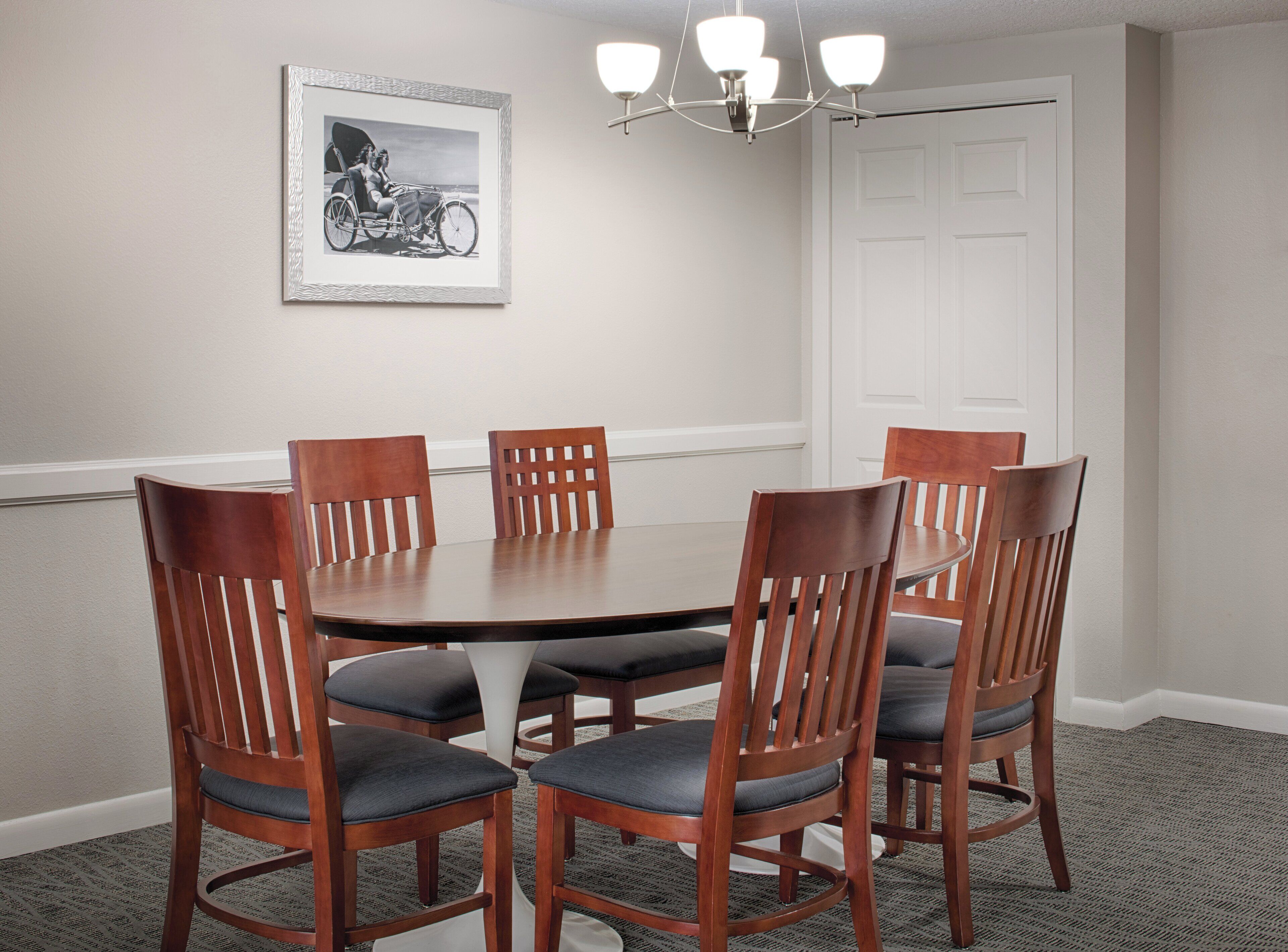 A conference room with a round table, six wooden chairs with cushioned seats, a framed black and white photograph on the wall, a white door, and a chandelier.
