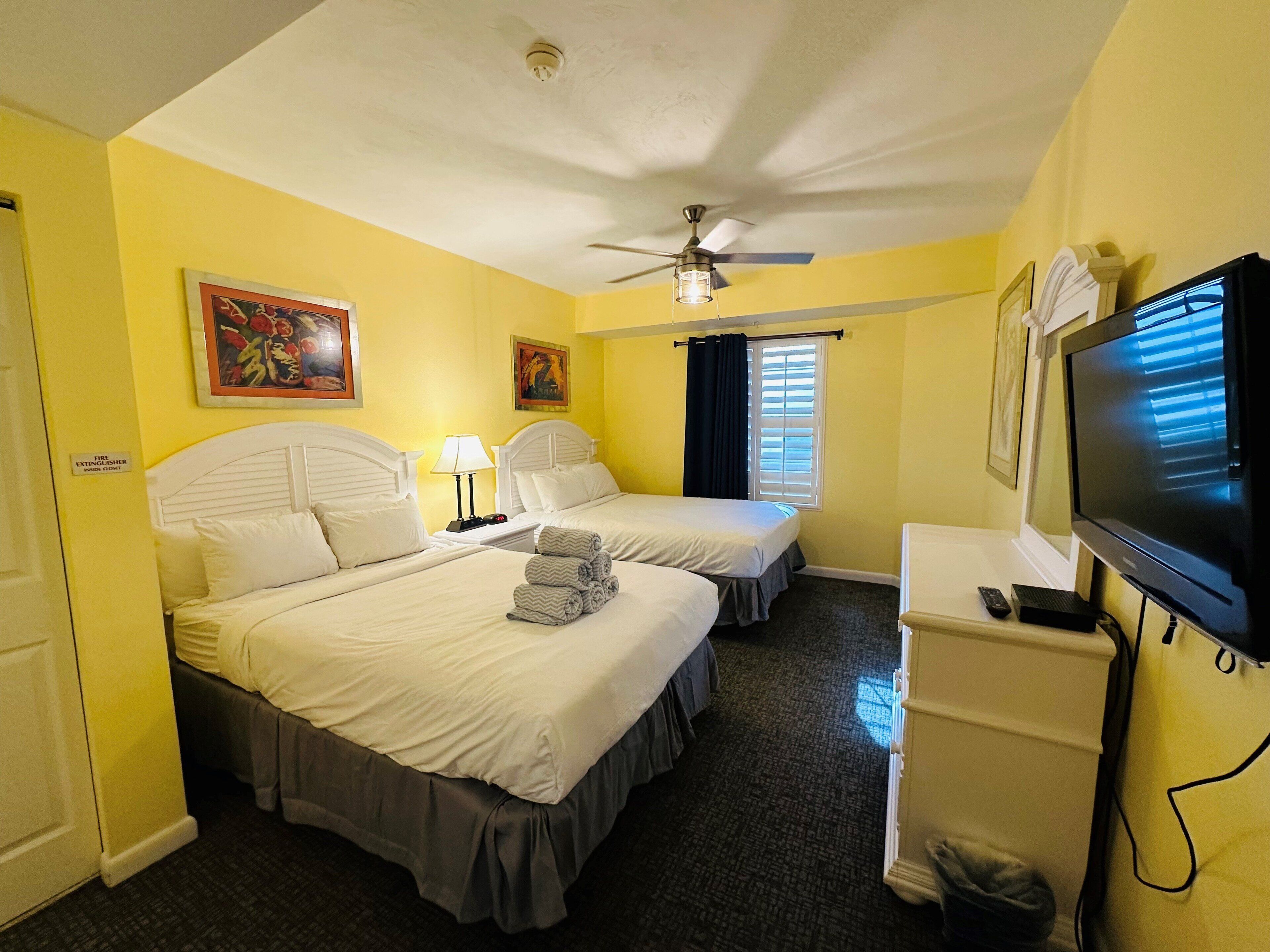 A hotel room with two beds, a ceiling fan, a television, and artwork on the walls.