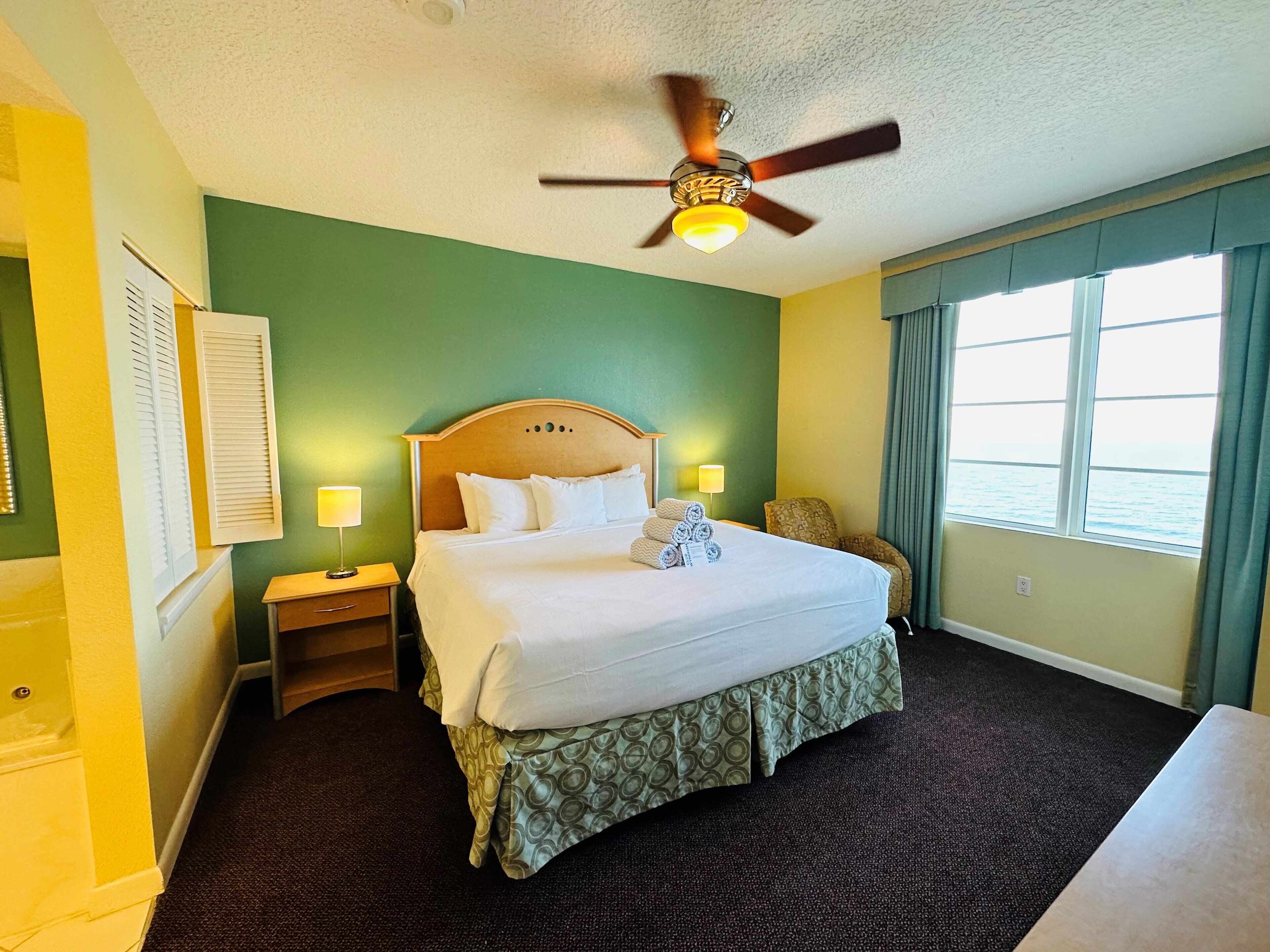 A hotel room with a bed, a ceiling fan, a window with curtains, and a nightstand with a lamp.