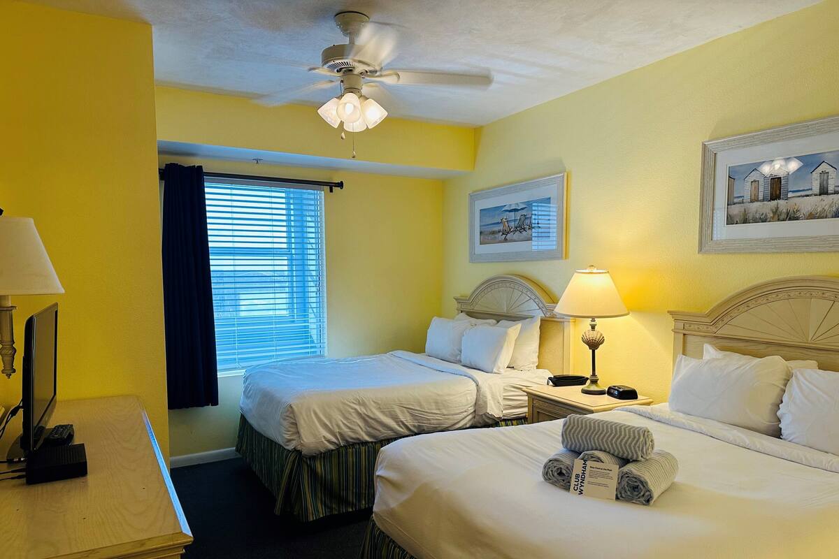 A hotel room with two beds, a ceiling fan, a desk with a lamp, and a window with blinds.