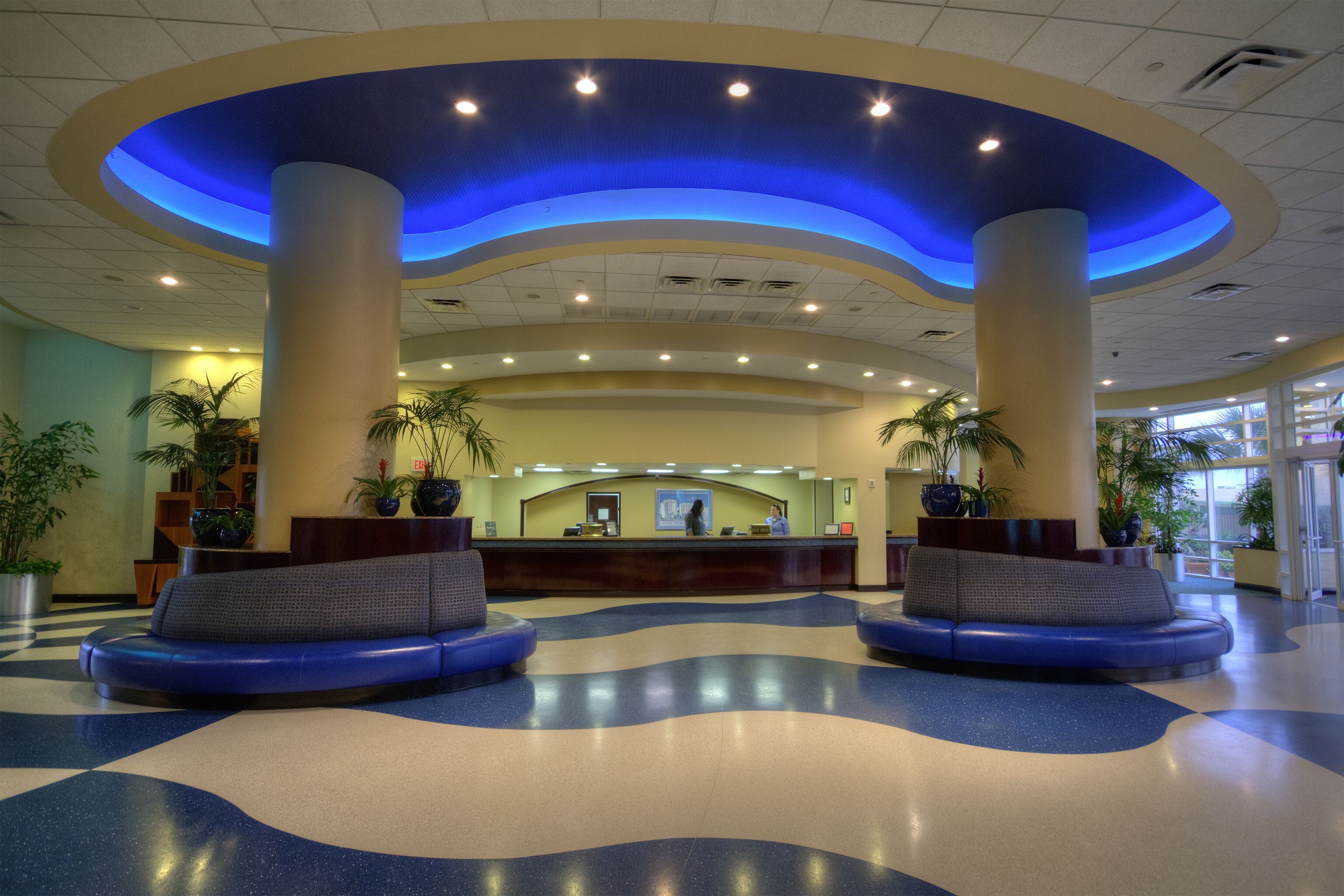 A lobby with a reception desk, two blue seating areas, and potted plants.