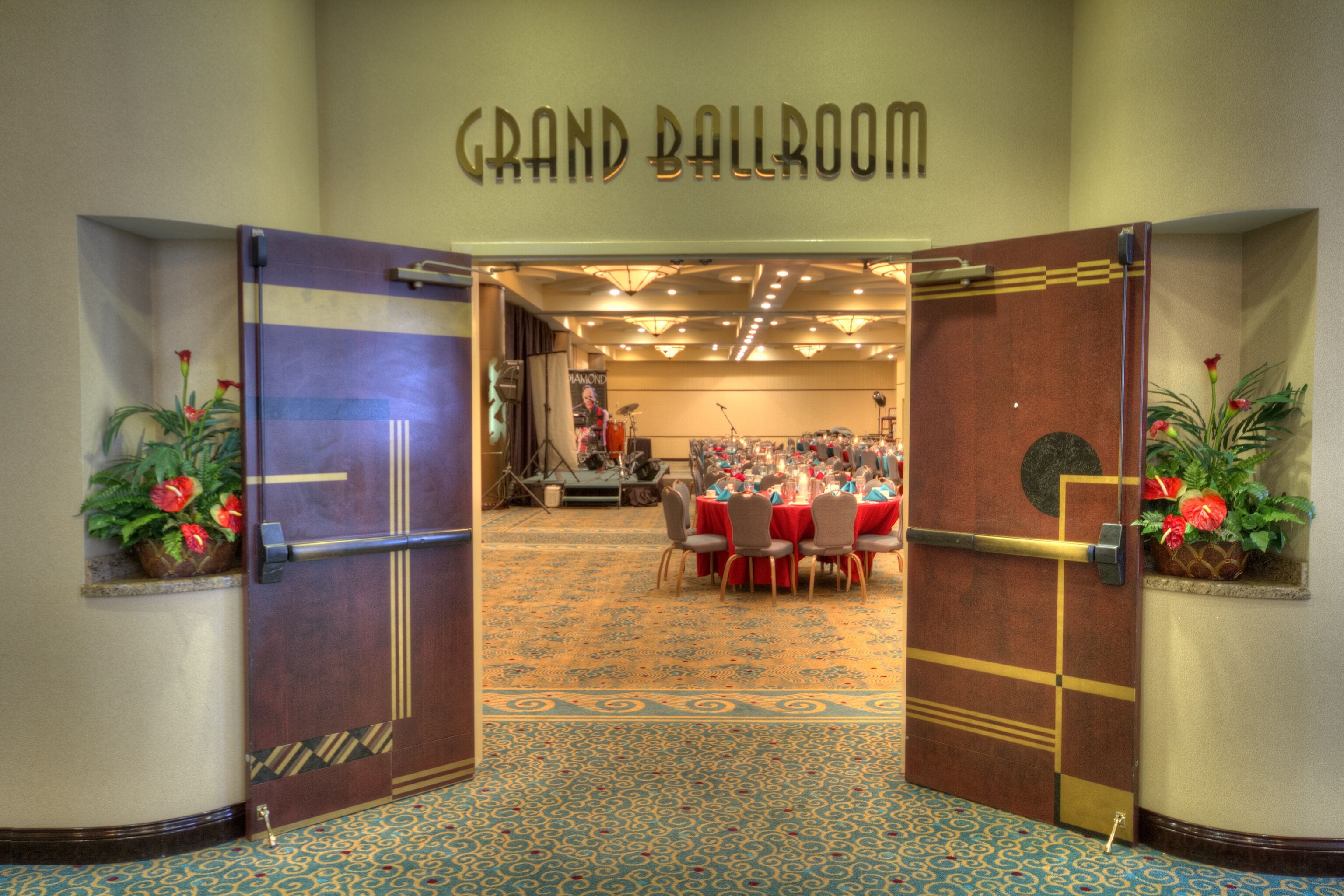 A grand ballroom with a patterned carpet, a stage with musical instruments, and red tables and chairs.