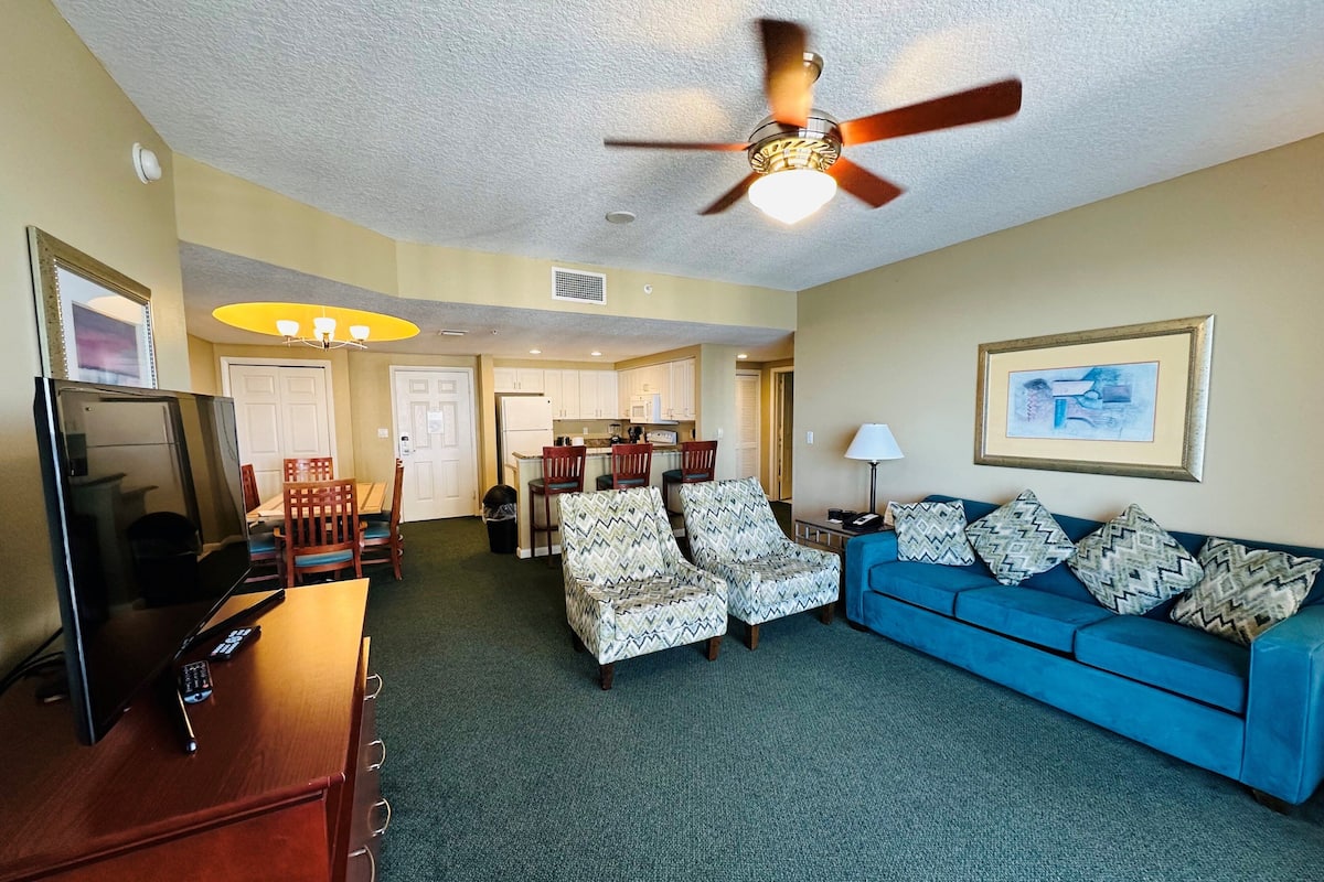 A living room with a blue sofa, patterned armchairs, a television, and a ceiling fan.