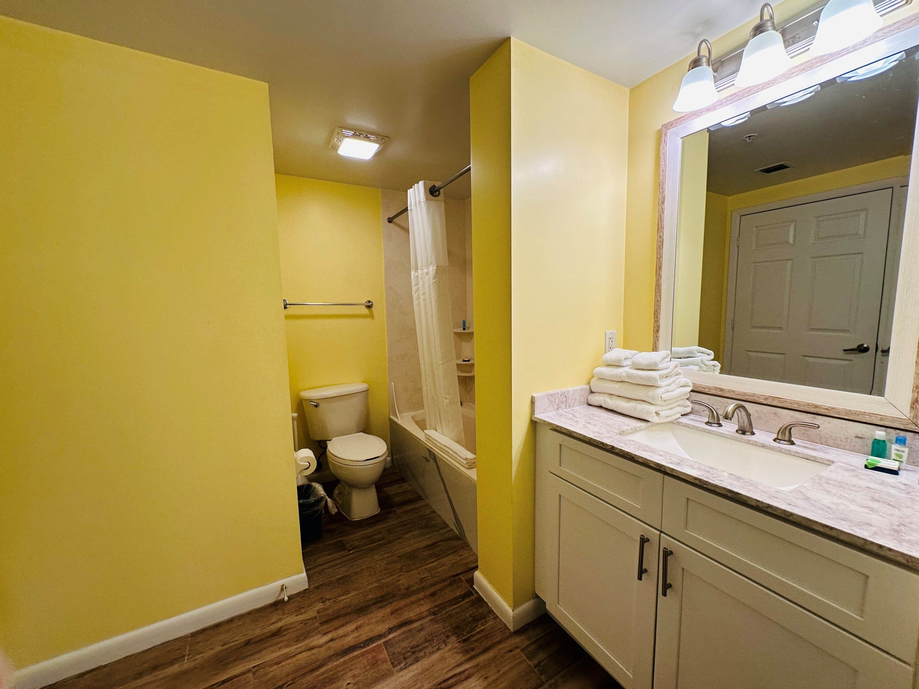 A bathroom with yellow walls, a wooden floor, a white toilet, a shower with a white curtain, a sink with a marble countertop, and a mirror above the sink.