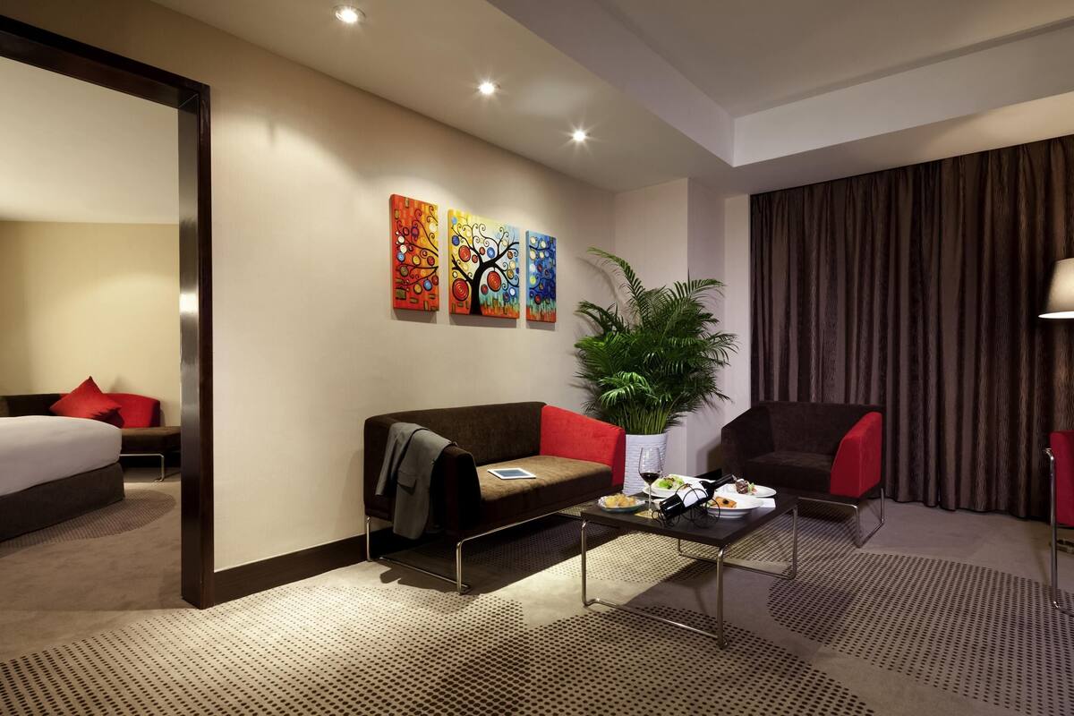 A hotel room with a bed, a sofa, a coffee table with a bottle and glasses, a plant, and three paintings on the wall.
