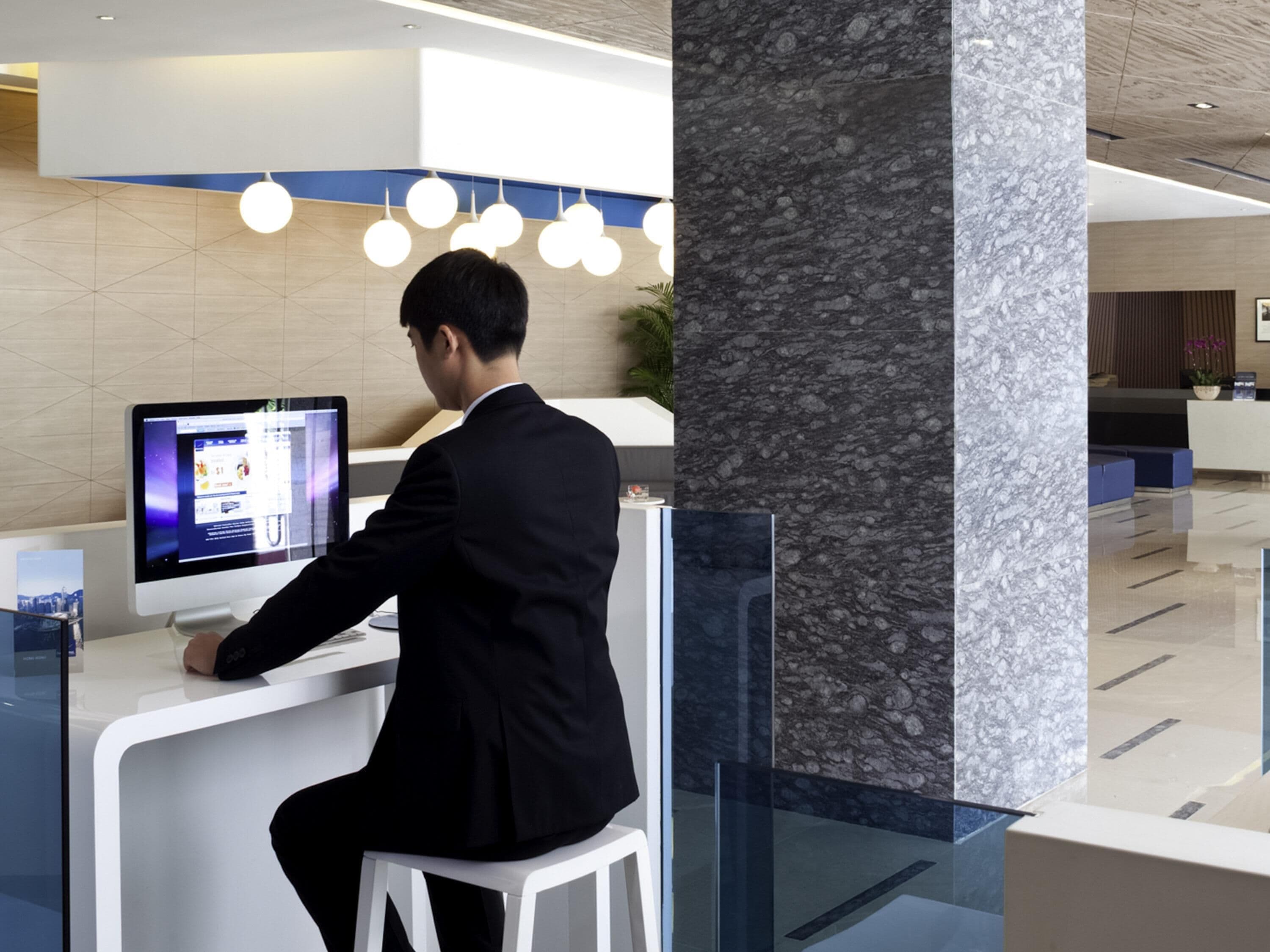 En mand sidder ved en reception med en computerskærm, i en moderne lobby med en marmorsøjle og et receptionsområde.