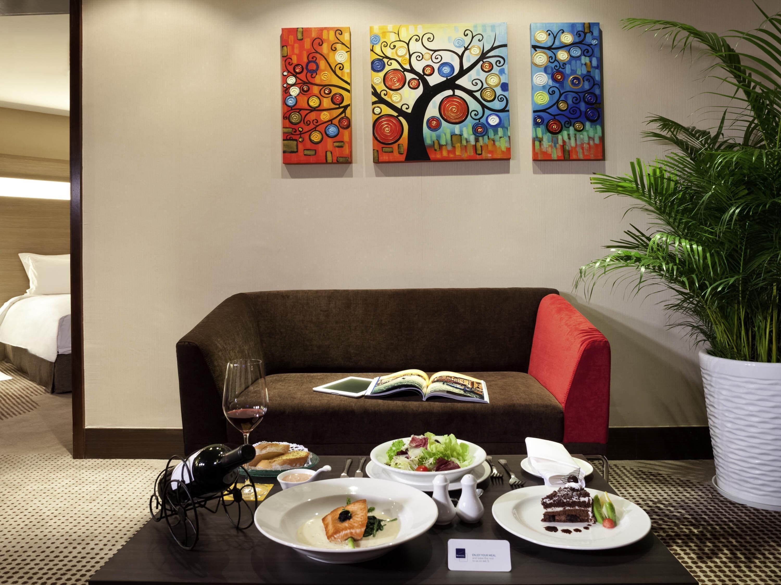 A hotel room with a brown sofa, a dining table with plates of food and a wine glass, and three colorful paintings on the wall.