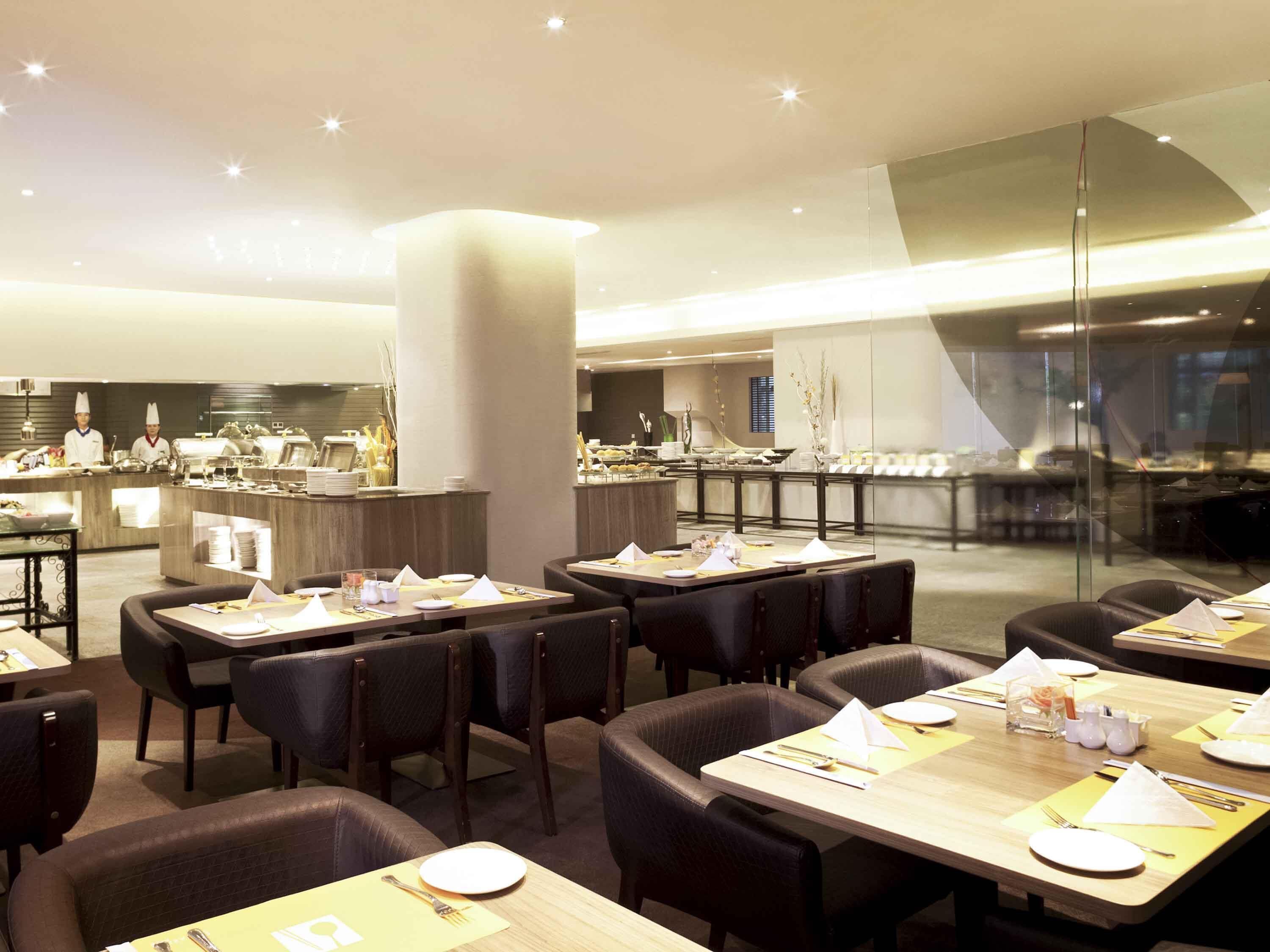Un restaurante moderno con varias mesas y sillas, una zona de cocina al fondo y un panel de vidrio que separa el área de comedor de la cocina.