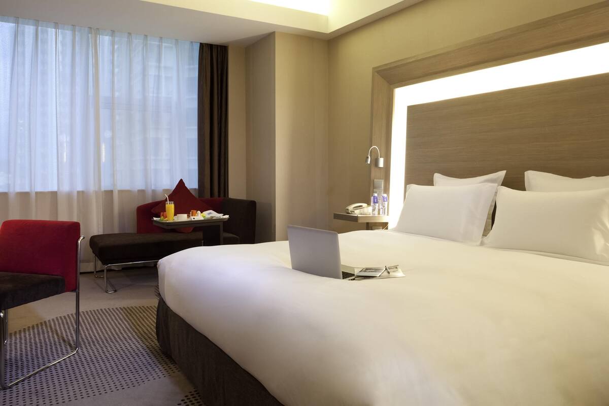 A hotel room with a large bed, a desk with a laptop, a chair, and a small table with a tray of breakfast items.