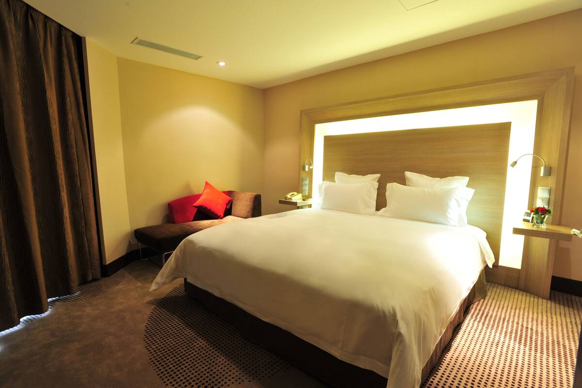 A hotel room with a large bed, a bedside table with a lamp, a chair with red cushions, and a large window providing natural light.