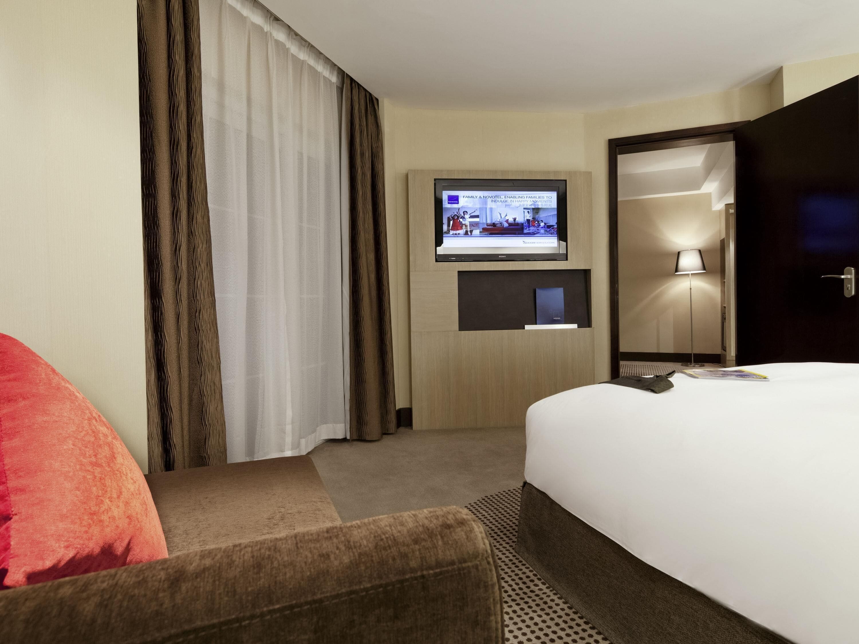 A hotel room with a bed, a TV mounted on the wall, a lamp, and a door.