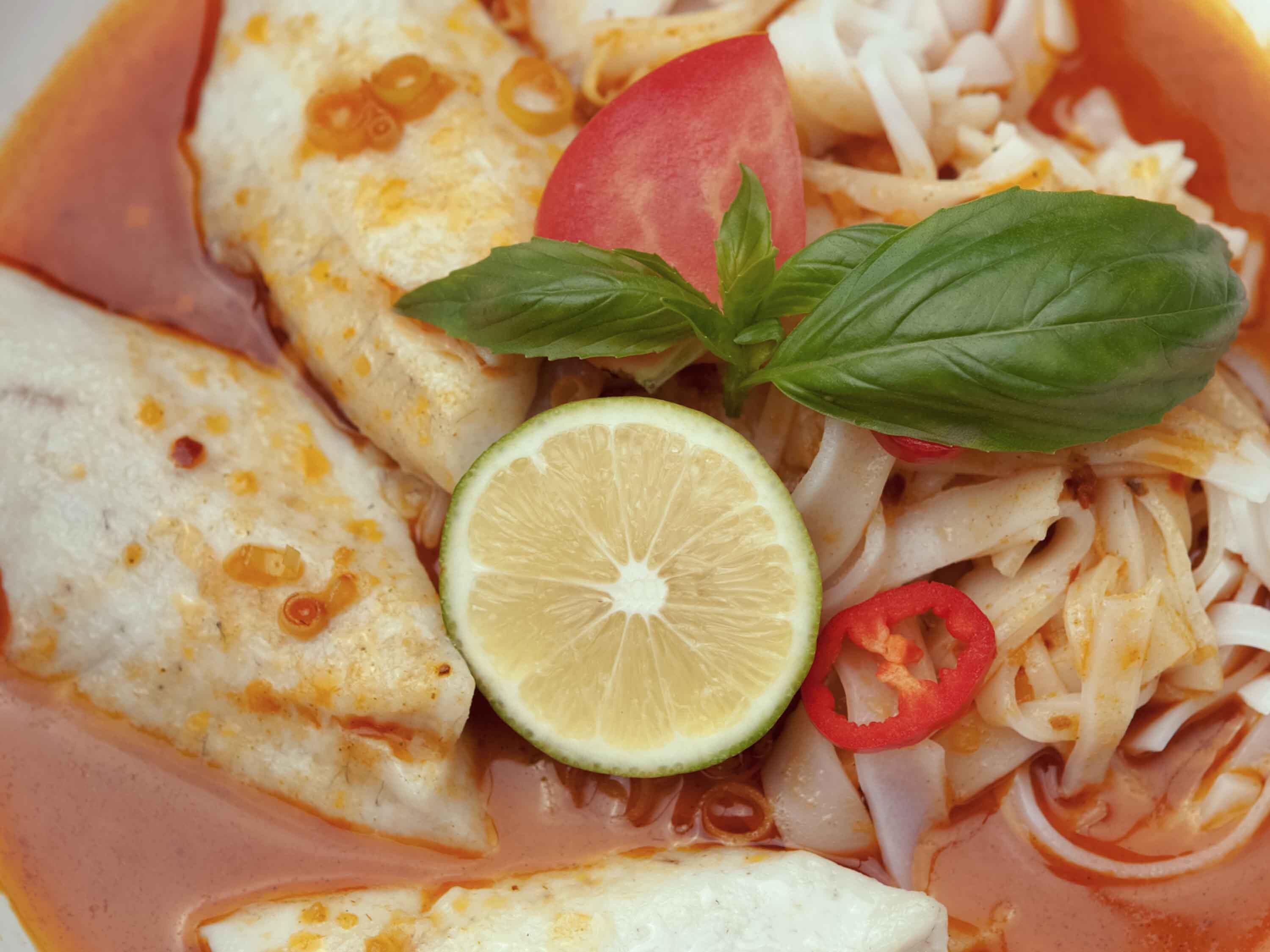 Un tazón de sopa de pescado con fideos, rodajas de tomate, un gajo de lima y hojas de albahaca.
