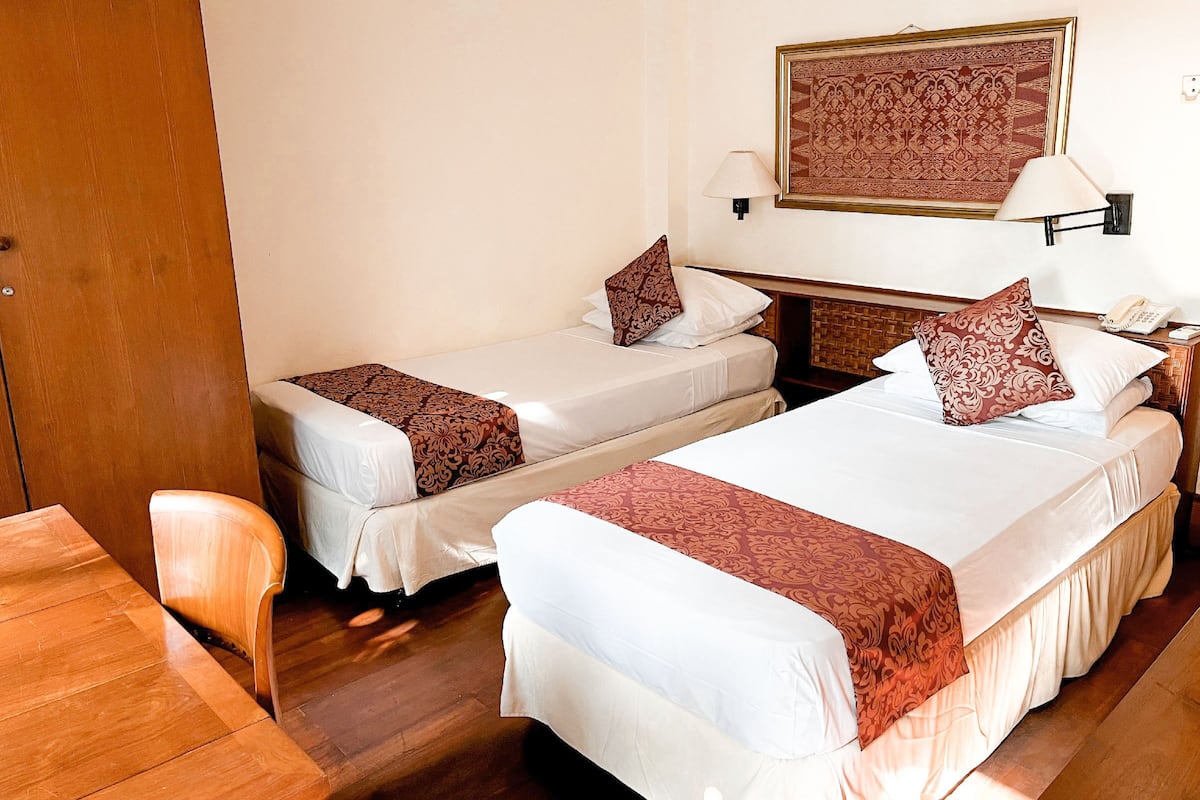 Dos camas individuales con sábanas blancas y almohadones marrones, un escritorio de madera y un teléfono instalado en la pared.